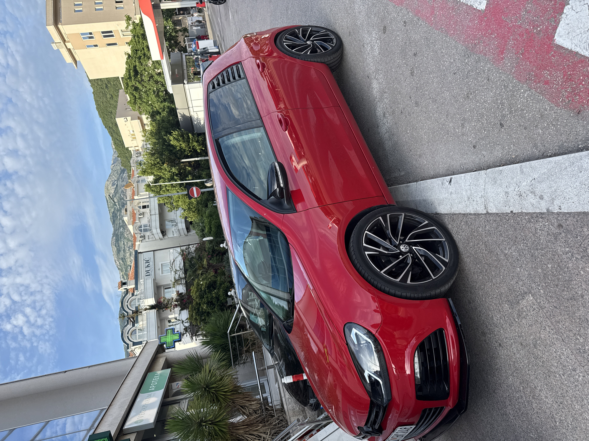 The image depicts an urban street scene in Budva, Montenegro, specifically identifiable by "VEGAPHARM APOTEKA" and "DUKIĆ APARTMANI" signage visible on buildings. The setting is daytime under a clear blue sky with scattered stratocumulus clouds.
In the foreground and mid-ground, a bright red Volkswagen Golf Mk6 hatchback is parked. The car features custom black multi-spoke alloy wheels with polished accents, low-profile tires, a black front lip spoiler, dark tinted windows, and black slatted louvers on the rear side windows. A partial license plate "BK 246" is visible on the front bumper, with "BK" likely indicating Budva registration. The vehicle appears clean and well-maintained. A dark-colored car is parked closely behind the red Golf, partially obscured.
To the left, a modern building with a grey facade and prominent glass windows houses "VEGAPHARM APOTEKA," indicated by a white sign with green lettering and a glowing green cross symbol. Several palm trees and green shrubs are planted along the building's base.
In the central background, various multi-story buildings line the street. A notable white building features arched windows and a sign reading "DUKIĆ APARTMANI." Another building with a beige facade is visible to its right. Lush green, tree-covered mountains rise in the distant background.
To the right, a gas station is visible with a distinctive red and white canopy structure covering fuel pumps. Indistinct human figures are visible near the gas station. A red and white "No Entry" road sign is positioned near the gas station entrance.
The ground consists of grey asphalt, with a white concrete curb adjacent to the red car's left side. Red and white painted markings are visible on the asphalt surface to the right of the vehicle. A red and white striped bollard is present between the two parked cars.
