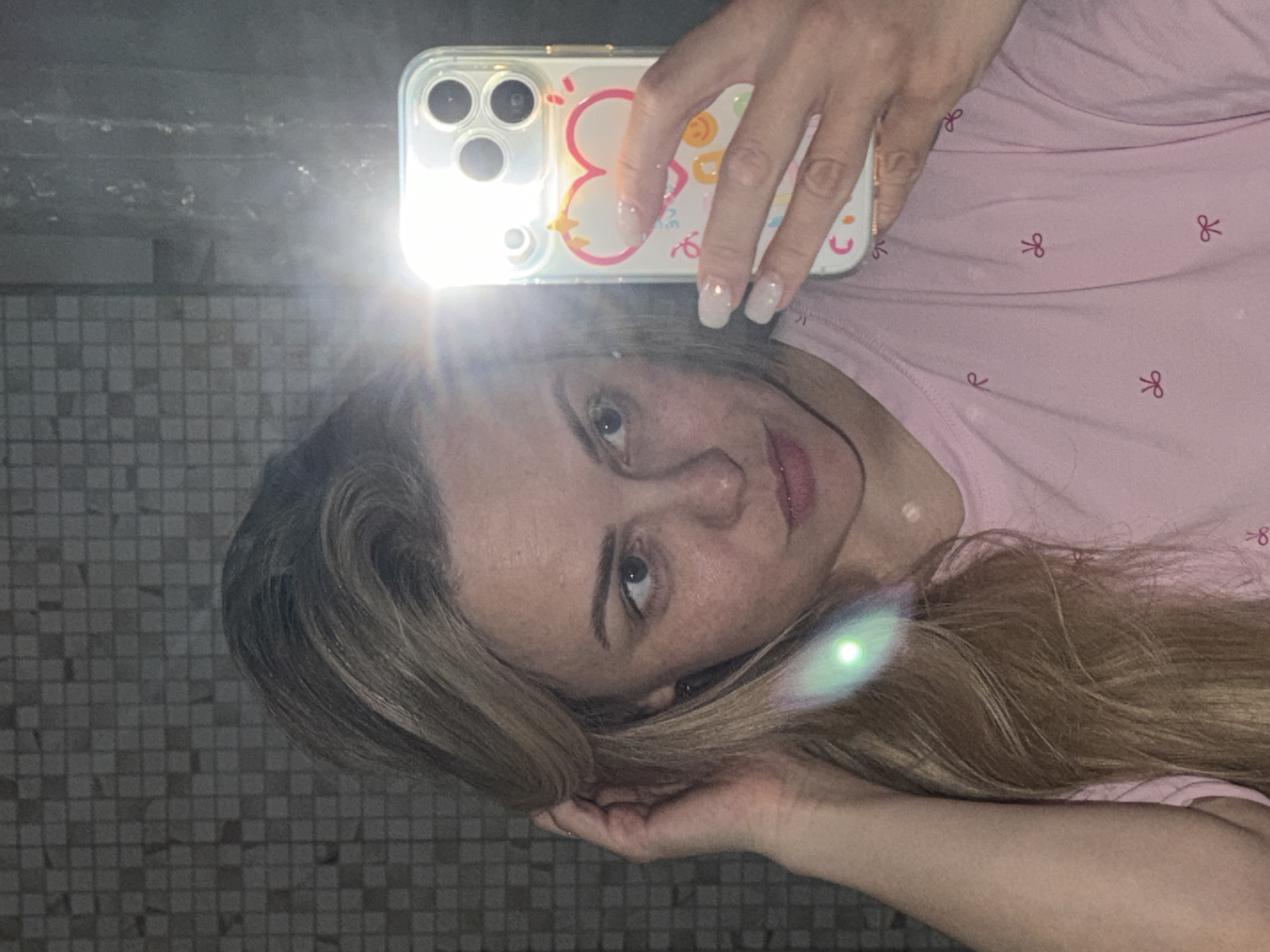 A woman is taking a selfie in a bathroom with a tiled wall. She is holding a smartphone with a clear case decorated with cartoon characters. She is wearing a pink shirt with small bow patterns. The location in the city of Votkinsk, Russia is not determinable from the image.