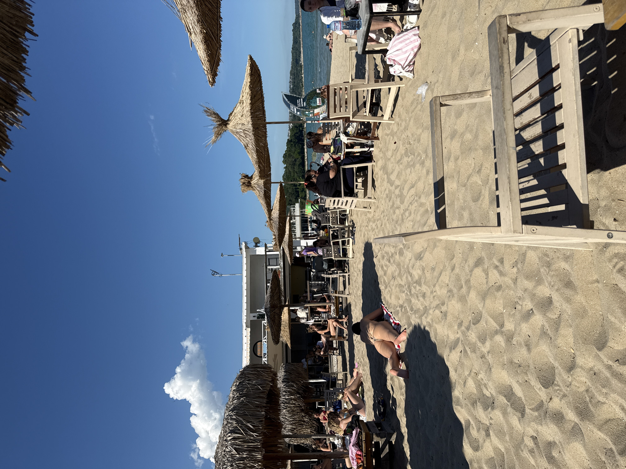The image depicts a sunny day at a beach in Varna, Bulgaria. In the foreground, a person is sunbathing on a towel, facing away from the camera. Several other individuals are seated at tables under thatched umbrellas, some eating or drinking. A white building with a balcony is visible in the background, alongside the beach umbrellas and chairs. The sea is calm, and the sky is clear blue with a few scattered clouds. The scene suggests a relaxed atmosphere at a beach resort.