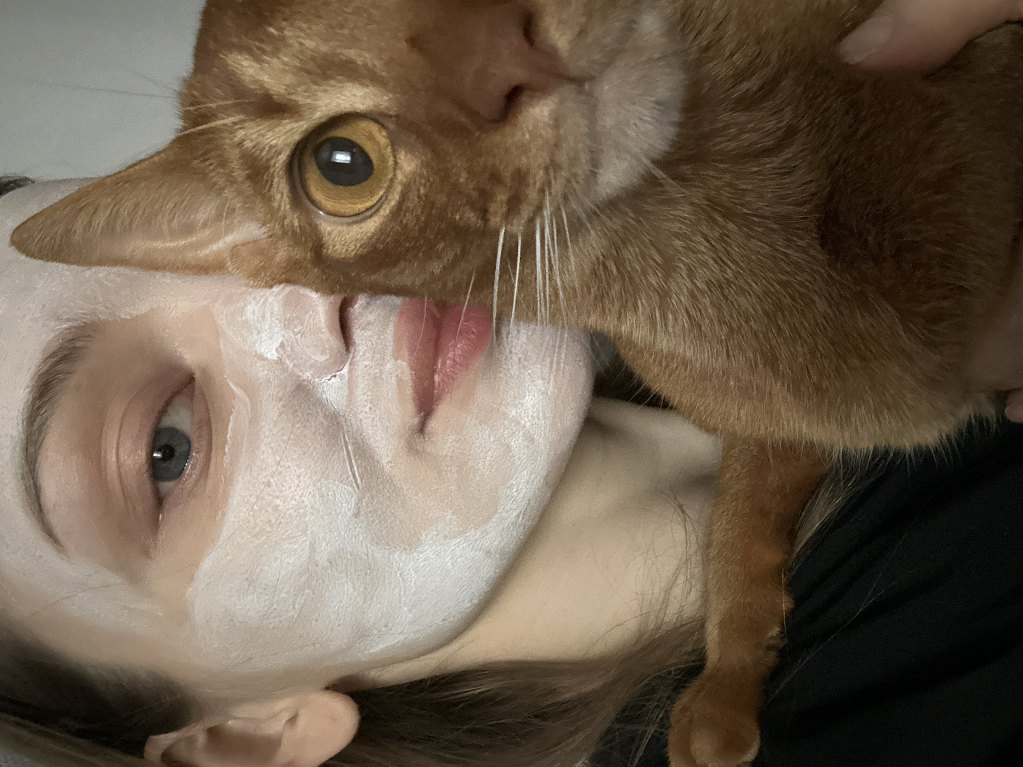A fair-skinned person lies on their back, face up, with a white facial mask applied. An orange tabby cat is positioned with its head and front paw resting against the person's face. The cat's eye is open and visible, as are its whiskers. The person's left eye is also open. The background appears to be a plain wall. The image was taken indoors. There is no definitive evidence to indicate that the photo was taken in Kazan, Russia.