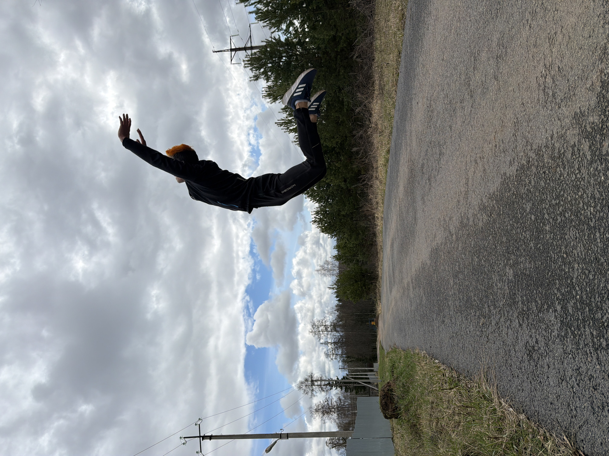 A person wearing dark clothing and Adidas shoes is airborne, oriented horizontally against a backdrop of a cloudy sky and a paved road. The person has orange hair and outstretched hands. The paved road is located to the right side of the image. A forested area lines the side of the road. Utility poles and wires are visible. Based on the presence of Russian words on the person’s pants and the landscape, it is likely this image was taken in the city of Chaikovsky, Russia.