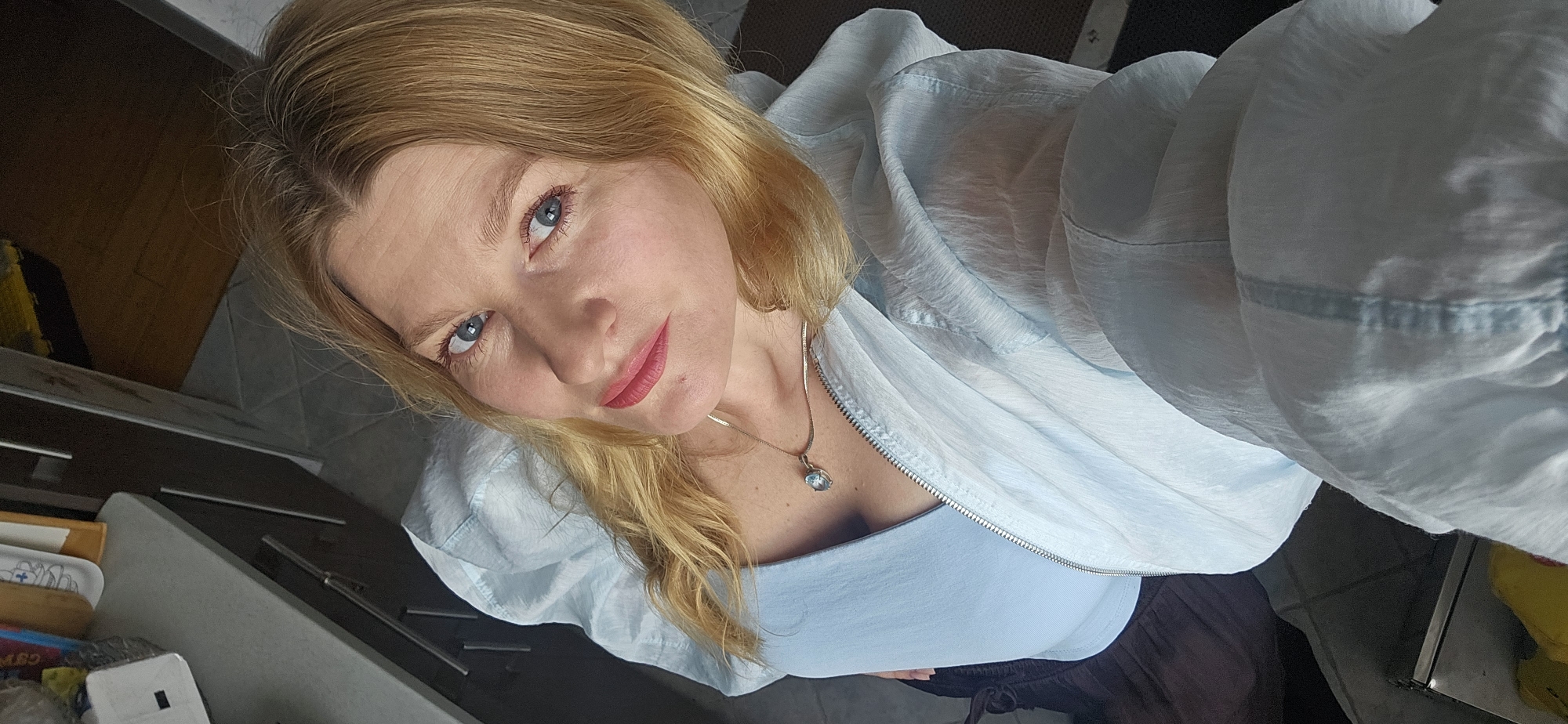 The image is a selfie taken from a high angle, looking down at a blonde woman with blue eyes and pink lipstick. She is wearing a light blue top and a light blue zippered jacket. A silver necklace with a light blue pendant is visible around her neck. The background appears to be an indoor setting, possibly a kitchen, with dark cabinets and a tiled floor. There are various objects on a countertop in the background, including containers and packaging. The overall lighting is natural, coming from an unseen source. The image does not contain any specific visual cues to identify the location as Krasnodar, Russia.