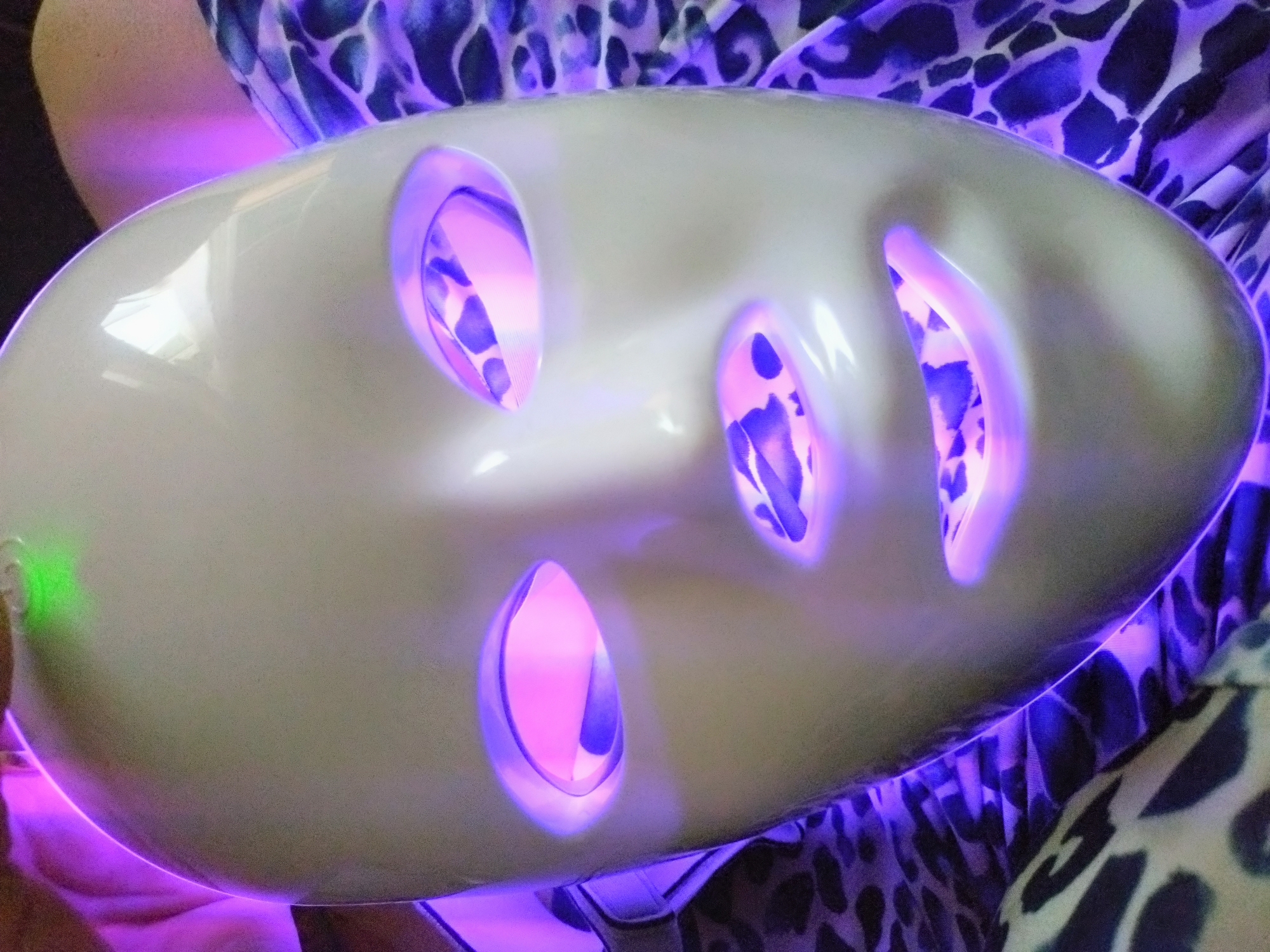 The image displays a close-up of a white, smooth, and elongated mask-like device, possibly a LED light therapy mask, held or worn by an individual. The mask features several oval-shaped cutouts that emit a strong purple-pink light. A small green light is also visible on the left side of the device.
Partial views of a person's shoulder and arm are visible, suggesting they are either operating or receiving treatment with the device. The person is wearing clothing with a distinctive purple and dark blue leopard print pattern, which is partially illuminated by the light emanating from the mask.
The scene takes place indoors, likely in a beauty salon, spa, or a private setting in Stara Zagora, Bulgaria. The activity depicted is the use or demonstration of this light-emitting device, suggesting a beauty treatment or therapeutic session. The dominant lighting comes from the device itself, casting purple hues on the surroundings and the person's clothing, creating an artificial, focused ambiance. No specific time of day or weather conditions are discernible, and there is no visible text in the image. The overall impression is one of modern skincare or wellness technology.