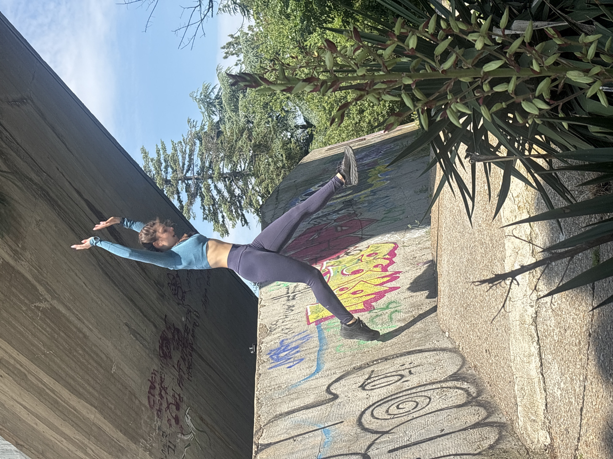 A young female, dressed in a teal long-sleeved cropped top and dark purple athletic leggings, is depicted mid-jump in an urban outdoor setting in Varna, Bulgaria. Her body is angled upwards and to the right, with arms raised above her head, palms facing forward, and legs bent, indicating a dynamic leap. She is wearing black athletic shoes. The background features a large, diagonal concrete bridge or overpass in the upper portion of the frame, showing textured surface details. Below the bridge, a concrete retaining wall is heavily adorned with various styles of colorful graffiti, including prominent yellow, blue, and black markings. Beyond the concrete structures, a clear blue sky with scattered white clouds is visible above a line of dense green trees. In the foreground, on the right, a tall plant with long, sword-like green leaves and a central stalk displaying numerous green and reddish buds occupies a significant portion of the frame. The scene is brightly illuminated by direct sunlight, producing sharp shadows on the concrete surfaces.