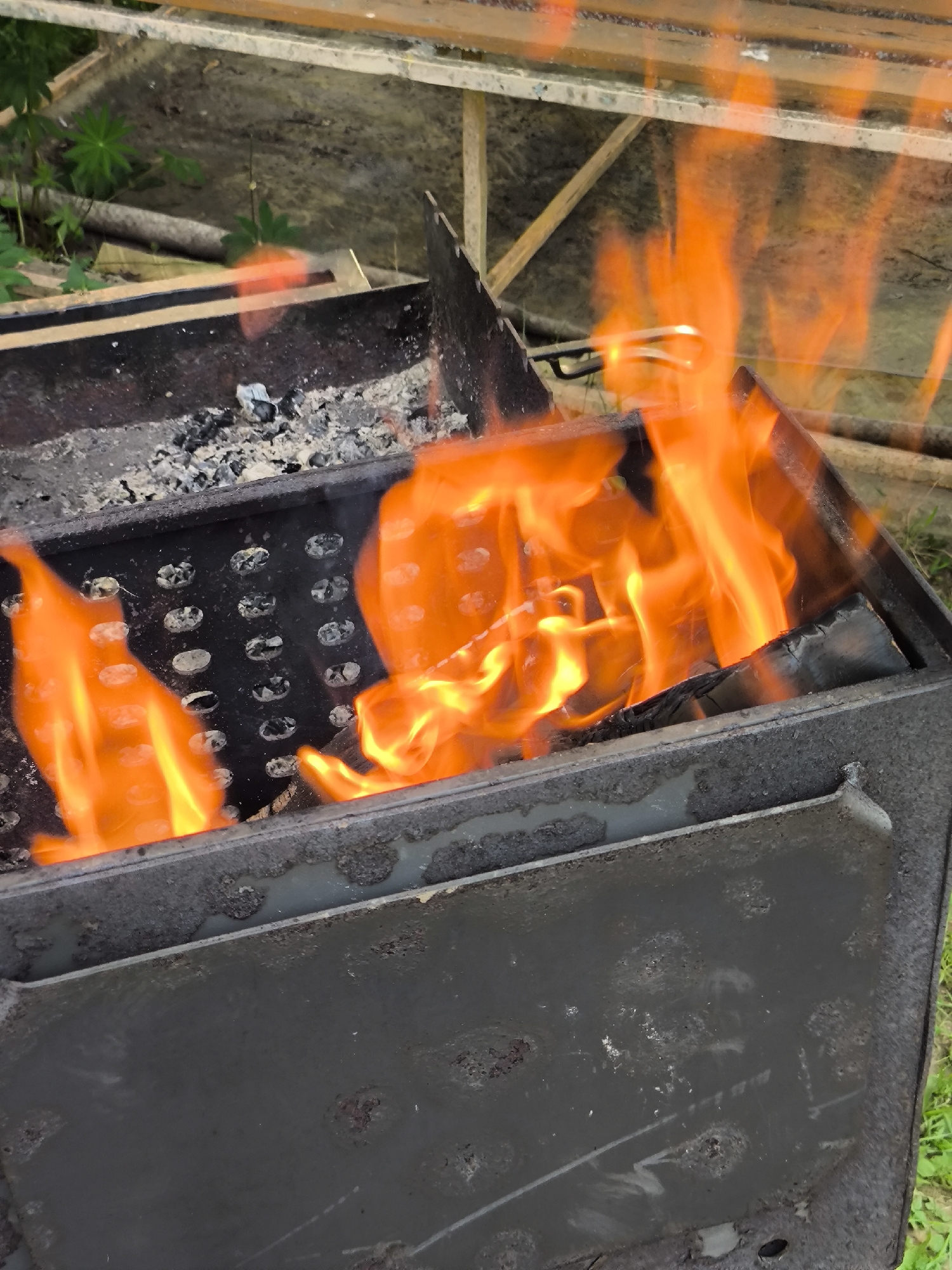 A close-up, eye-level shot shows a metal barbecue grill with bright orange flames engulfing pieces of wood and charcoal. The grill is dark and appears worn with some rust. In the background, the blurry shapes of green foliage and wooden planks can be seen, suggesting an outdoor setting. The lighting is bright, highlighting the intensity of the fire.