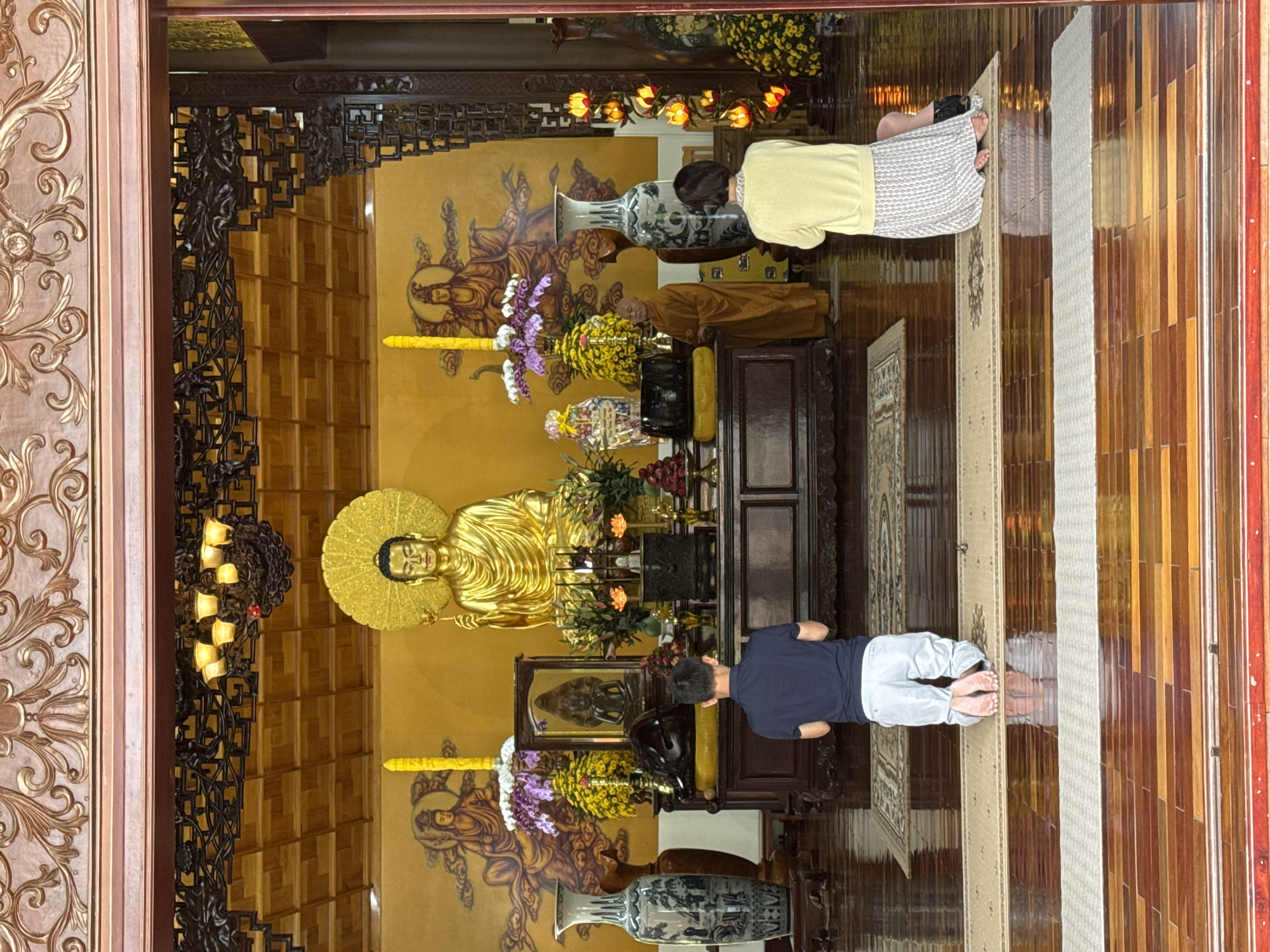 The photo captures the interior of a temple in Da Lat, Vietnam. A golden Buddha statue takes center stage, positioned before a backdrop featuring painted figures. An altar is arrayed with flowers, fruit, and other religious objects. Two individuals are seen kneeling in prayer, one dressed in light-colored clothing and the other in dark blue. A monk in saffron robes stands to the side. The temple's architecture is characterized by ornate woodwork and decorative details. A carpet is partially visible on the polished wood floor.