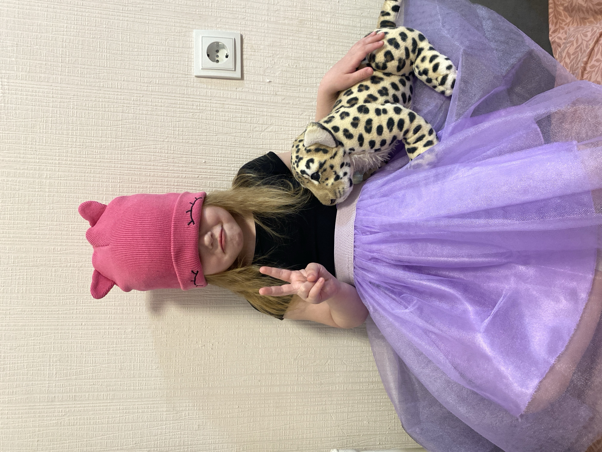 A young girl is pictured indoors, likely in a home in Izhevsk, Russia, judging by the electrical outlet on the wall. She wears a pink knit cap with stitched-on cat ears and a black top. The girl is also wearing a purple tulle skirt with a light purple waistband. She is holding a stuffed cheetah toy with her right hand. The background includes a textured beige wall. The girl is holding up two fingers in a peace sign.