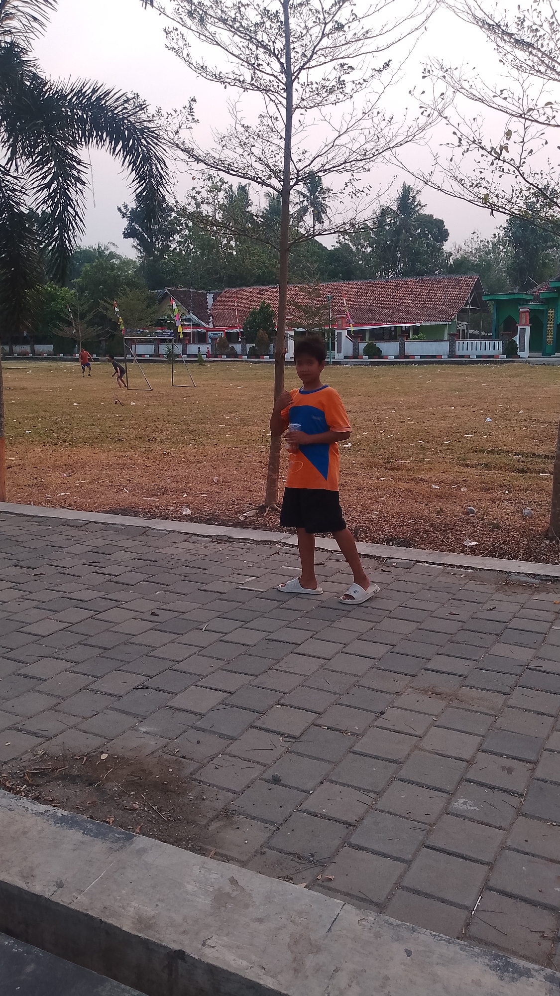 A young boy stands on a paved walkway in Karanglangu, Indonesia. He is wearing an orange and blue t-shirt and black shorts, with white sandals on his feet. He holds a clear plastic cup in his left hand. The walkway is made of square gray pavers and a concrete curb is visible in the foreground. Behind him, a grassy field stretches out, with a few scattered leaves and twigs. In the middle ground, buildings with red tile roofs and white walls are visible, along with some trees and a soccer goal. A palm tree is on the left side of the image. The sky is overcast.
