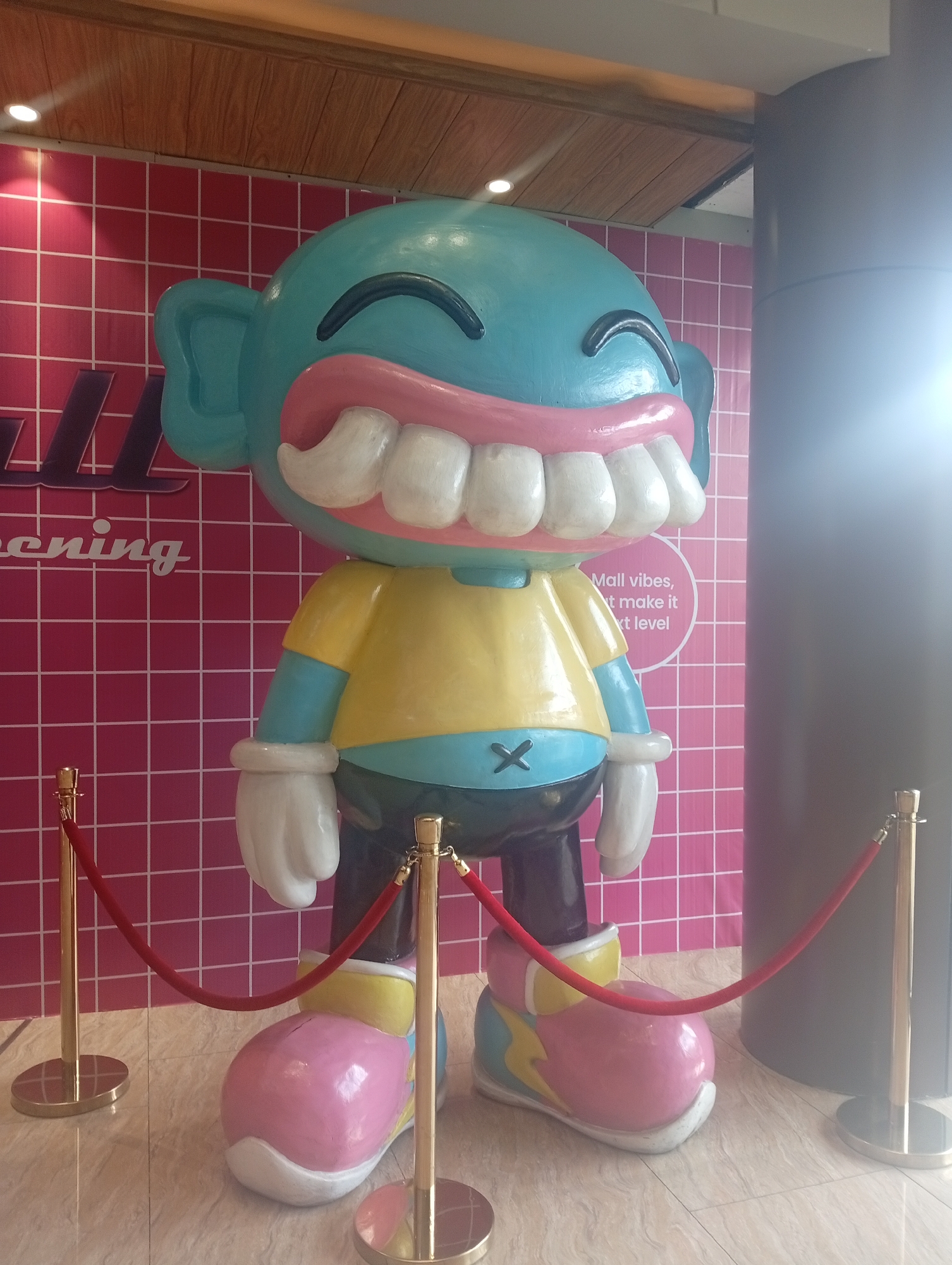 A brightly colored statue stands indoors on a light-colored, reflective floor in Denpasar, Indonesia. The statue is humanoid, featuring a blue head with large, cartoonish white teeth, a yellow shirt, black pants with an "X" on the waist, white gloves, and pink shoes with yellow and blue accents. A red velvet rope barrier held by two gold stanchions encircles the statue. The background is a wall with a pink and white grid pattern. Text that includes the word "screening" is faintly visible on the left side of the wall. A circular column stands to the right of the statue. The top of the wall has a wood paneling.