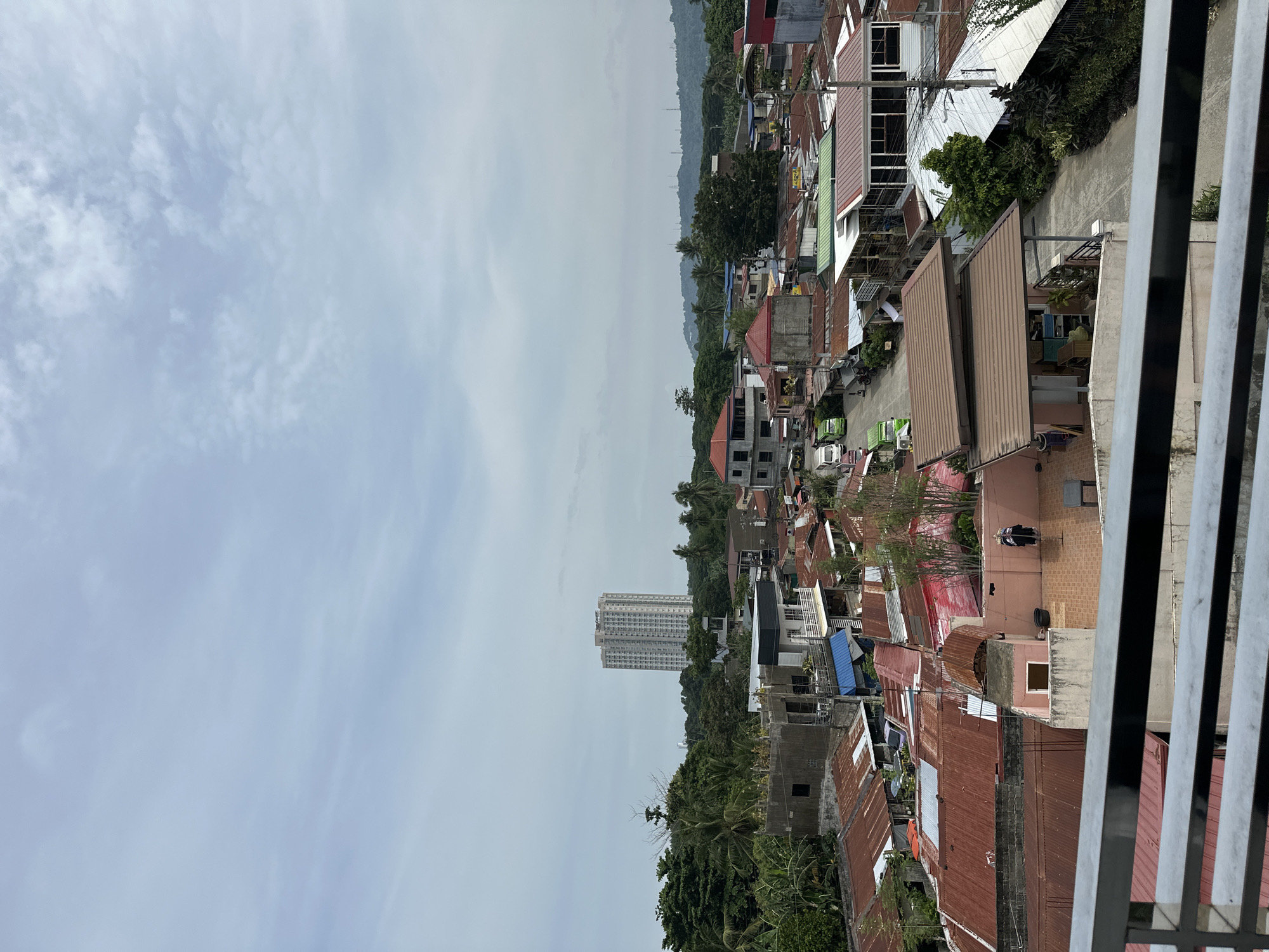 A cityscape in Davao City, Philippines, shows a mix of residential and commercial buildings. Numerous houses with rusty corrugated iron roofs fill the lower foreground, while a tall, modern apartment building rises in the middle distance. The sky is overcast with scattered clouds. A metallic railing is in the extreme right foreground.