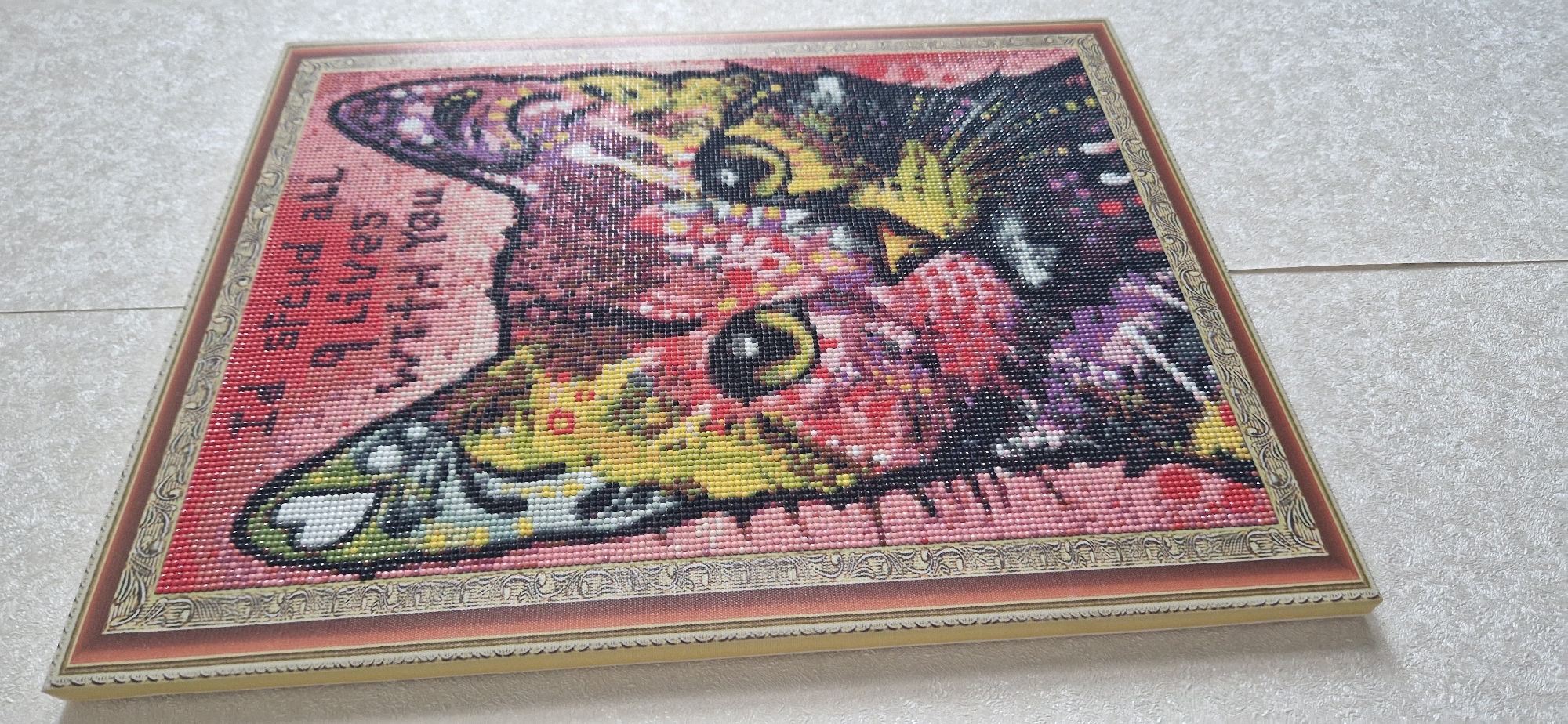 A diamond painting of a cat's face is framed and hanging on a textured wall. The cat is depicted with a colorful, abstract design, featuring a prominent pink and red background. Above the cat's head, text reads "I'd spend all 9 lives with you." The painting is in a ornate frame with gold and reddish-brown detailing. The wall has a light-colored, textured finish, possibly wallpaper. There are no people or animals present in the image. The location is unknown.