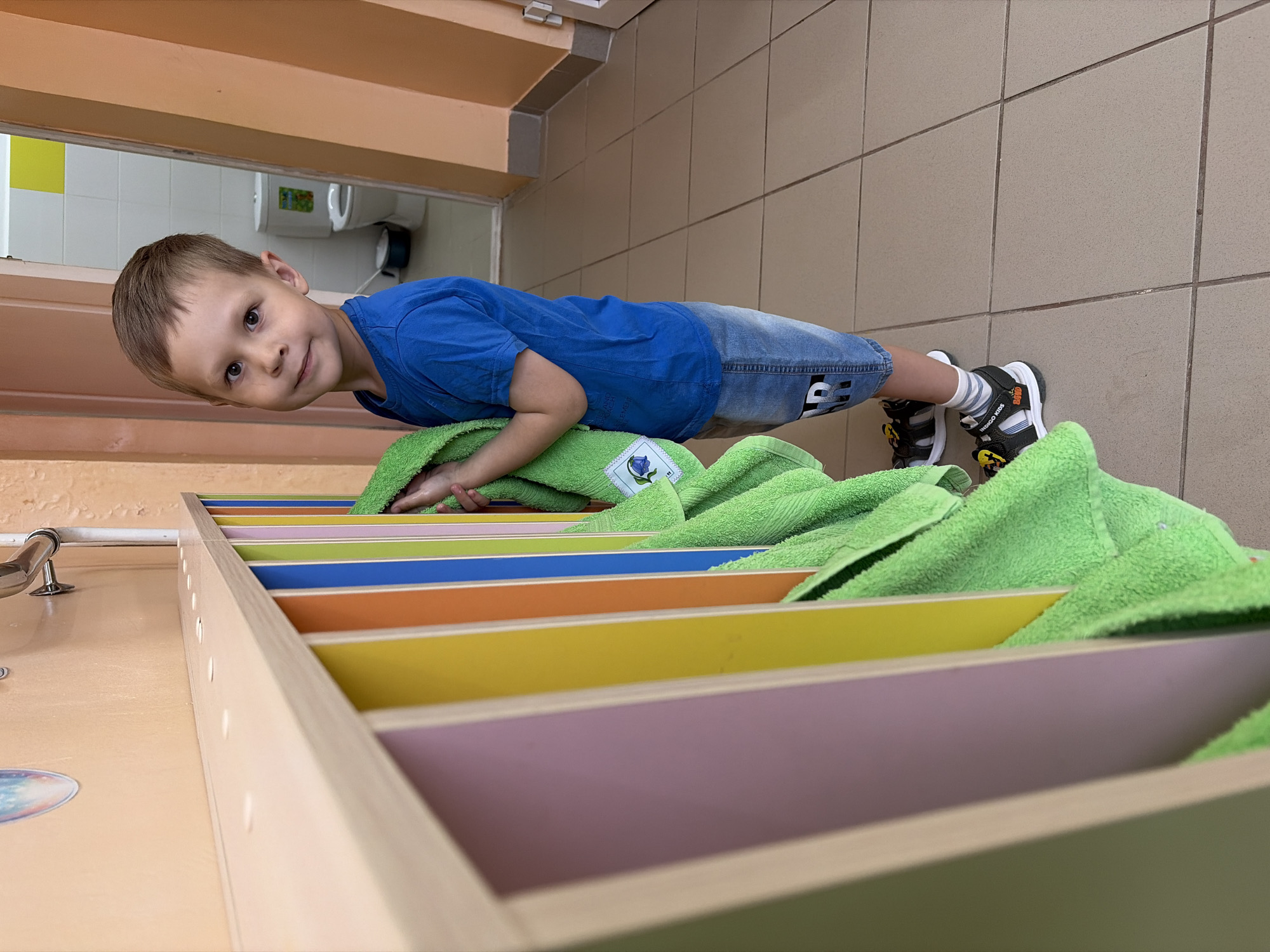 A young boy is doing push-ups on a green towel. The towel is placed on top of a multi-colored shelf with rows of pink, yellow, orange, blue, green, and light green. The boy is wearing a blue shirt and jean shorts with black and white shoes and gray socks. The location appears to be a bathroom. The background shows a beige-tiled wall. The image was taken in the city of Chaikovsky, Russia.