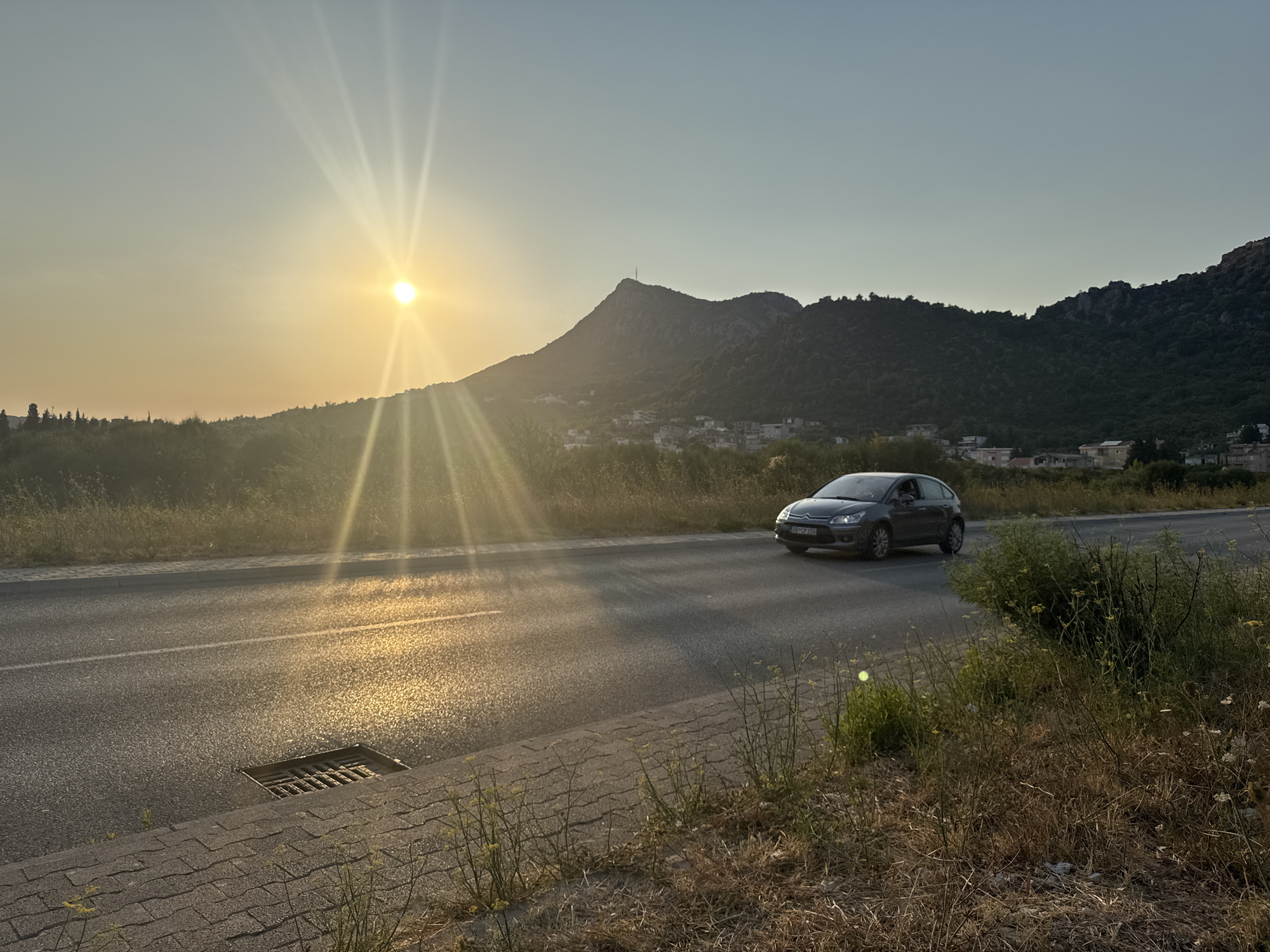 An outdoor, horizontal, eye-level, medium shot captures a scenic view of a road next to a body of water, with a mountain and a small town in the background. The sun is low in the sky, casting a golden glow over the scene. The road is asphalt with a white line down the middle. On the right side of the road, there is a car driving away from the camera. The car is dark gray and appears to be a hatchback. In the background, there is a mountain with a small town nestled at its base. The town has white buildings with red roofs. The sky is clear and blue. The overall impression is one of peace and tranquility.