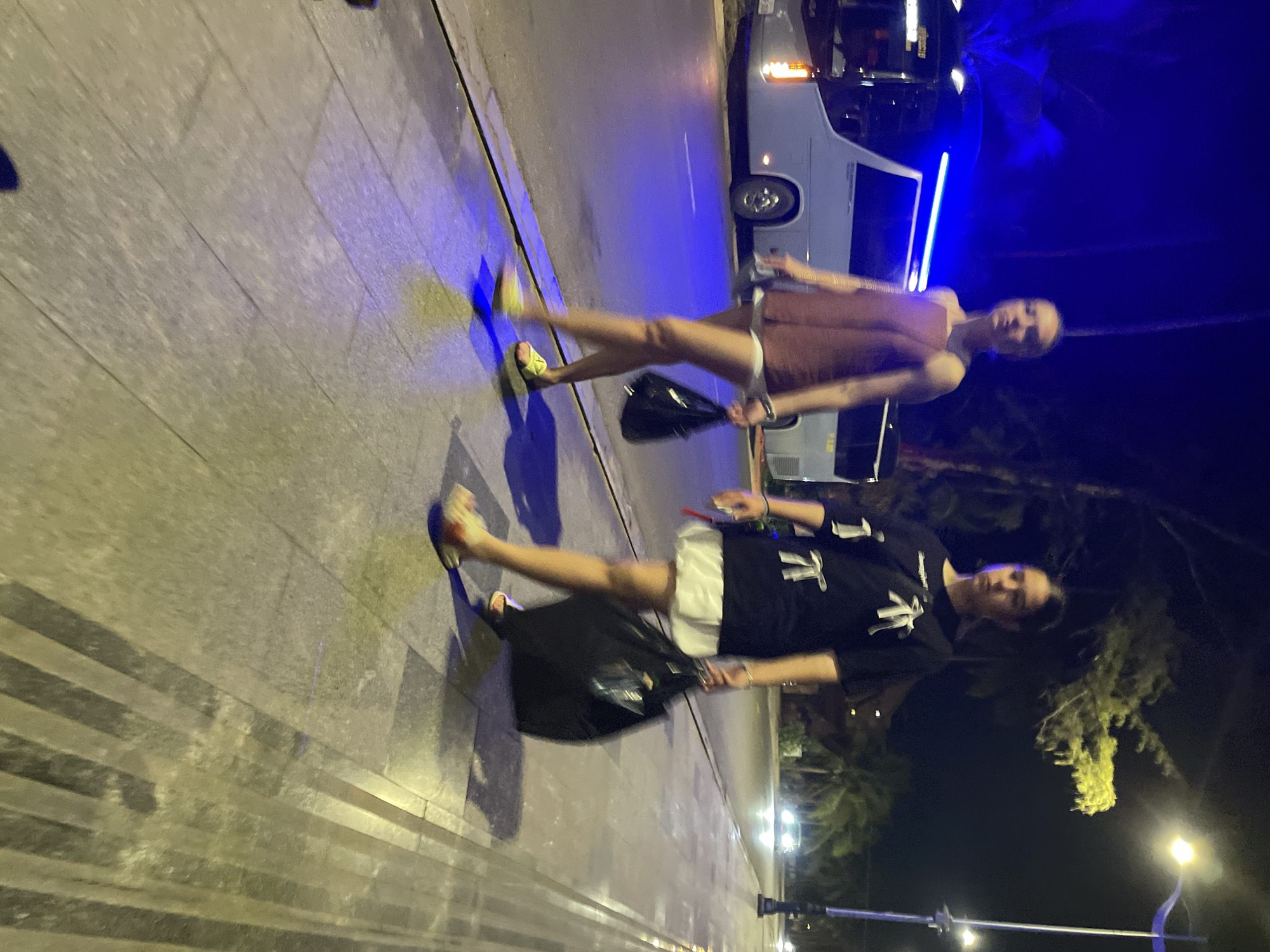 Two young women walk on a sidewalk at night in Phu Thuy Ward, Vietnam. The woman on the left wears a sleeveless, reddish-brown top and white shorts, carrying a black bag in her right hand and yellow slides on her feet. The woman on the right wears a black t-shirt with white graphics and a white skirt, carrying a large black bag in her right hand and sandals on her feet. A bus is parked on the left side of the road, illuminated by blue lights. Streetlights cast yellow and blue light on the sidewalk and road. Palm trees are visible in the background.