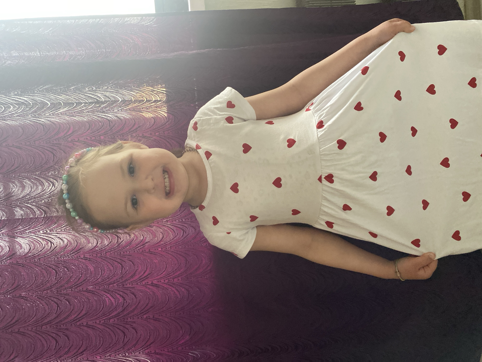 A young girl lies on a bed. She is wearing a white dress with red heart patterns, and a beaded necklace and headband. She is smiling. The bed has a purple patterned bedspread. The image was taken indoors, possibly in Chaikovsky, Russia, though the location is not definitively confirmed by the image content.