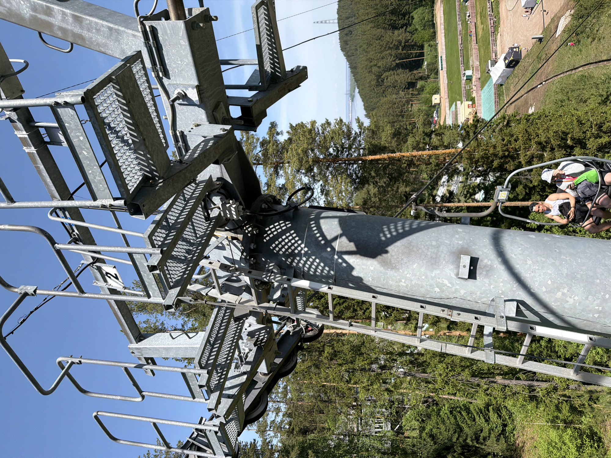 The image shows a view from a chairlift ascending a ski slope near Прикамский, Russia, on a sunny day. The gray metal structure of the chairlift tower is in the foreground, featuring a ladder, walkways with perforated metal surfaces, and support beams. Beyond the structure, green trees and vegetation cover the slope. At the top right, a ski jump or a landing area is visible. The background includes glimpses of a river or lake and a clear sky. Two people are seated in a chairlift ascending toward the upper part of the lift.