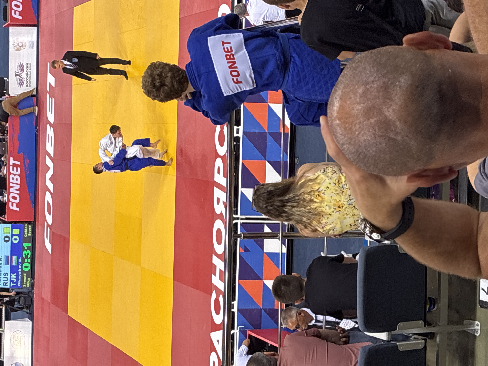 The image captures a judo match in progress, viewed from a spectator's perspective. In the foreground, the back of a bald man's head is visible, with his arm extended upwards, potentially interacting with another person or the railing. He is wearing a dark watch on his wrist. To his left, a man in a black t-shirt is seated, facing away from the camera. To the left of the bald man, another spectator in a pink t-shirt is seated, partially visible.
In the mid-ground, a judo match is taking place on a yellow and red mat. Two male judokas, one in a blue gi and the other in a white gi, are engaged in a grapple. A referee in a dark suit stands nearby, observing the match. In the background, a large scoreboard displays match information, including "TJK," "RUS," "0 0," and a timer showing "0:31." The word "FONBET" is prominently displayed on a banner above the scoreboard and on the back of a judoka's uniform in the mid-ground. Spectators are seated in tiered seating behind the mat. The environment appears to be an indoor sports arena.