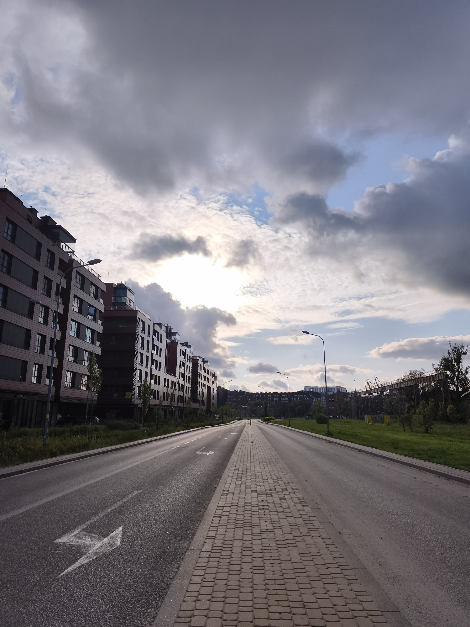 The image captures a street scene in the city of Gdańsk, Poland. In the foreground, a paved road runs into the distance, with a distinct white arrow painted on it, indicating a turn. Adjacent to the road is a pedestrian walkway made of light-colored cobblestones. On the left side of the road, a row of modern apartment buildings with dark facades and numerous windows stretches along the street. Trees and grass line the sidewalk in front of the buildings. In the background, more buildings and infrastructure, including what appears to be an industrial conveyor belt system, are visible. The sky is filled with dramatic, heavy clouds, with patches of sunlight breaking through, suggesting a dynamic weather condition. There are no people or vehicles visible in the frame.