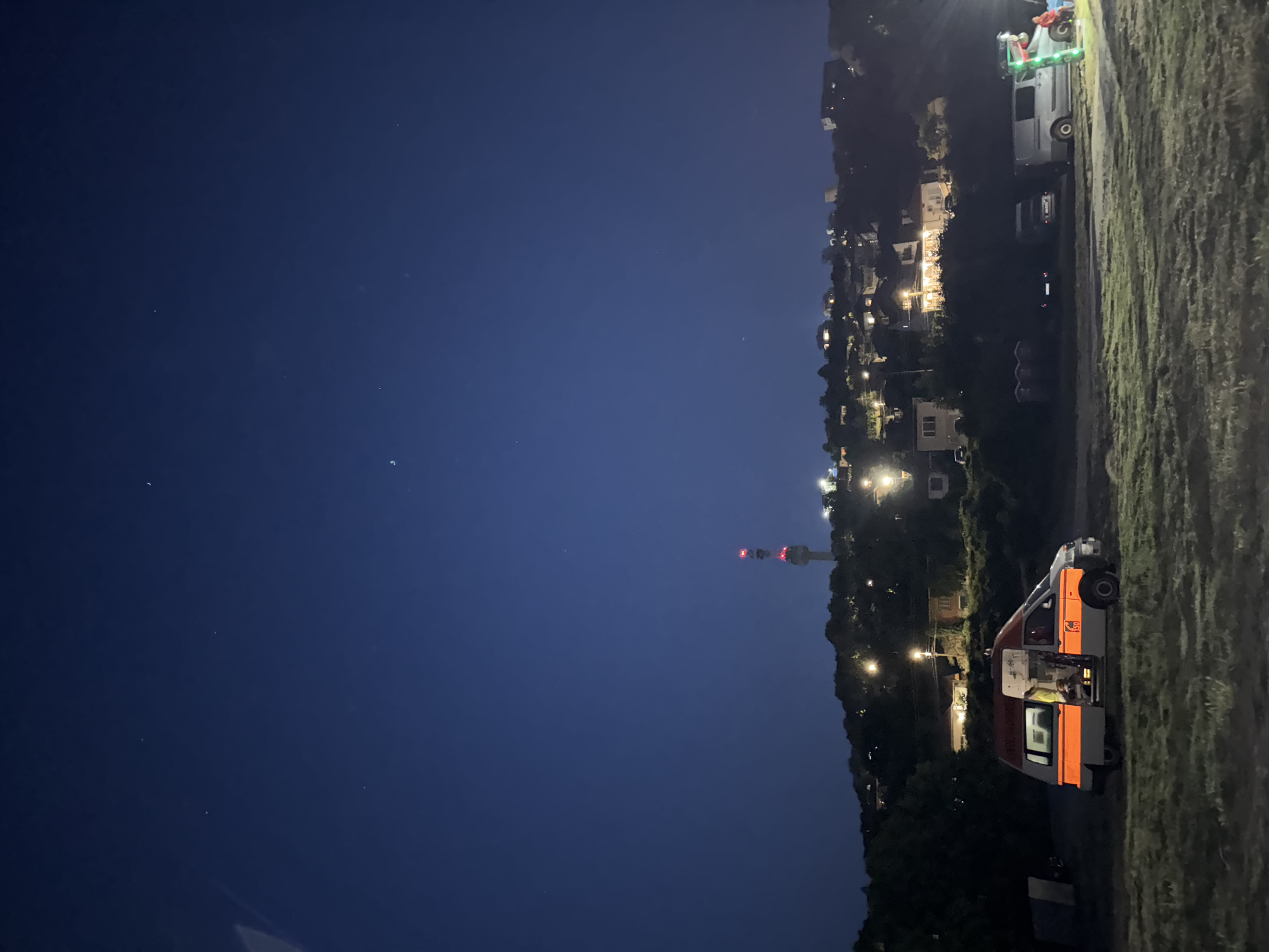This image captures a nighttime scene in Tutrakan, Bulgaria. The sky is a deep, clear blue, indicating a cloudless night, with several stars visible. In the foreground, on the right side of the image, a grassy embankment slopes down.
Dominating the mid-ground is a row of buildings and structures on a hillside, illuminated by various lights, suggesting a populated area. A prominent feature on the hillside is a tall tower with red and white lights, likely a telecommunications or observation tower.
Further to the right, parked on a dirt or gravel path near the embankment, are several vehicles. One is a white van with orange accents and visible lights, with its door open and a person inside wearing a yellow vest. Another vehicle, a dark SUV, is parked behind it. Further up the embankment, another white van is visible, adorned with green lights. The presence of these vehicles and the open door of one van suggest some form of activity or camping, possibly related to an event or a service.
The overall mood is serene, with the lights of the town and the vehicles creating a warm contrast against the dark night sky. The image appears to have been taken at night, likely after sunset. No visible text is present in the image.