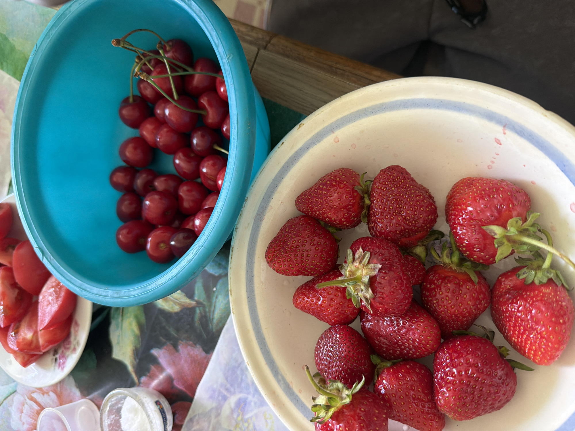 This image, taken outdoors at a private residence in Dnipro, Ukraine, depicts a close-up of a table set for a meal. In the foreground, a white bowl filled with fresh strawberries is visible. To the right, a bright blue bowl overflows with ripe cherries. Behind these bowls, a plate of sliced tomatoes rests on a floral-patterned tablecloth. A small glass container of salt is also present on the table. The setting suggests a casual outdoor gathering.