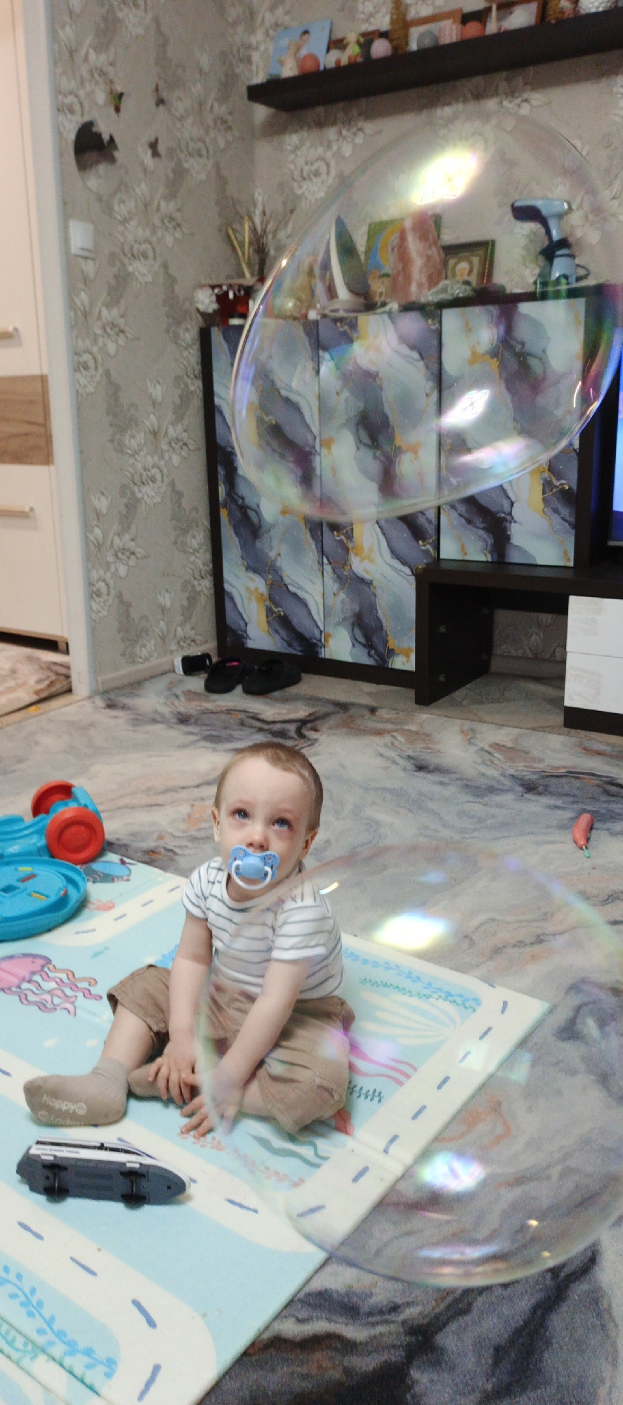A young child sits on a play mat in a room. The child has blond hair, blue eyes, and a pacifier in their mouth, wearing a striped shirt and khaki pants. The play mat has a light blue background with a jellyfish illustration. A toy car sits on the mat near the child. A large soap bubble floats in the air in front of and above the child. In the background, there is a television stand with decorative items on top and a shelf mounted on the wall. Shoes are near the base of the television stand. The wallpaper has a floral pattern. The location is Chaikovsky, Russia.