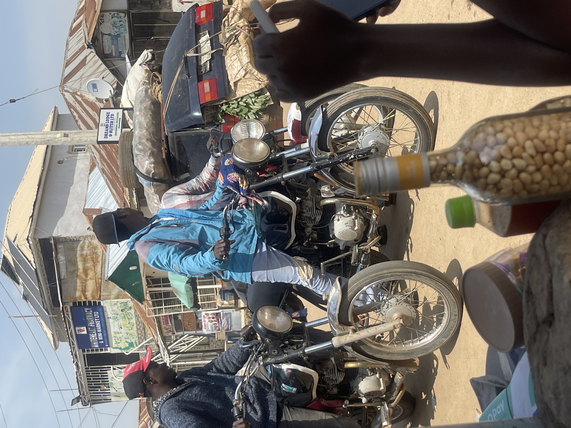 This image captures a bustling street scene in Abuja, Nigeria, likely during the daytime given the strong shadows and bright sunlight. Two individuals are prominently featured, both appearing to be on or near motorcycles.
The foreground is dominated by a motorcycle with a blue jacket draped over its handlebars. A person wearing a blue jacket and white pants is partially visible, seemingly interacting with the motorcycle. Behind them, another motorcycle is visible. To the right, a clear bottle filled with what appears to be small nuts or seeds sits on a surface, possibly a table or another motorcycle. The background reveals a typical urban Nigerian setting with corrugated metal roofs, buildings, and various signs.
One sign indicates "WHITEHEART PHARMACY & MINI-MARKET LTD," suggesting a commercial area. Another sign reads "DREAMS LODGE & SUITES LTD," indicating nearby accommodation. The presence of multiple motorcycles implies a common mode of transportation and potential for delivery services in the area. The overall atmosphere seems to be one of everyday commerce and activity, with the people appearing focused on their tasks. The sunlight suggests it's a clear, possibly warm day.