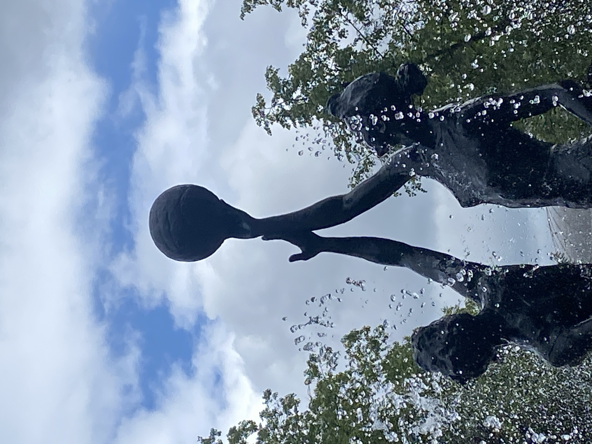 The image shows a low-angle view of a dark-colored sculpture fountain set against a cloudy sky and tree foliage. The sculpture appears to depict figures holding a round object, possibly a ball. Water droplets are visible, indicating the fountain is active. The photograph does not offer enough information to identify the specific location in Заволжье, Russia.