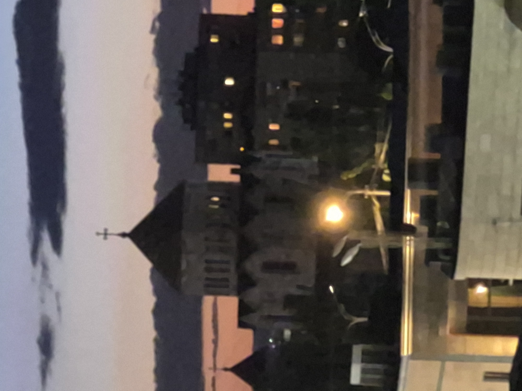 The image, correctly oriented, displays a twilight cityscape in Yerevan, Armenia. A prominent stone church or cathedral with a pointed roof and a cross on its spire occupies the left-center of the frame. Its facade shows architectural details and some illuminated windows. To the right of the church, a bright street lamp emits a warm, yellow glow. Further to the right and in the background, a tall, dark building, possibly an apartment block or office building, has numerous windows lit from within.
The sky, visible above the structures, transitions from a light blue-grey at the top to a warm orange and pink hue near the horizon, indicative of dusk or early evening. Dark cloud formations are present in the sky. The foreground also includes dimly visible elements, likely parts of other buildings or structures. No people or visible text are discernible in the image. The scene conveys a calm, urban atmosphere as day transitions to night.