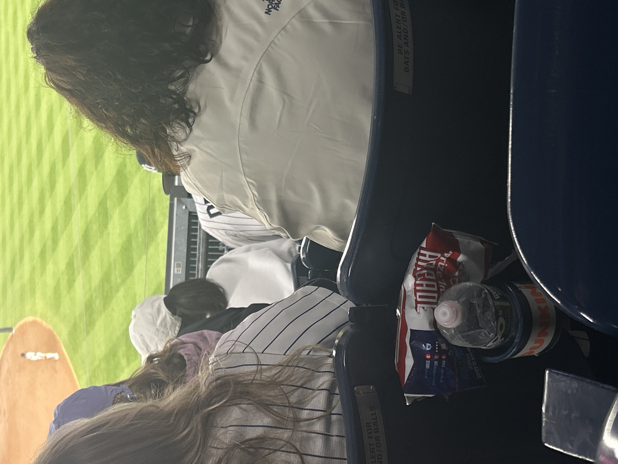 The image shows a partial view of a baseball game from stadium seating. A group of spectators is seated, including a woman with long brown hair wearing a striped shirt and another person wearing a white North Face jacket. A Dunkin' coffee cup, a water bottle, and a bag of "Arcade" chips are in the seat's cup holder. A sign that reads "BE ALERT FOR BATS AND/OR BALLS" is attached to the seat. The backdrop suggests a baseball field with green grass. Given the striped shirts, baseball game, and the brand of the coffee, the location is likely Yankee Stadium in New York City.