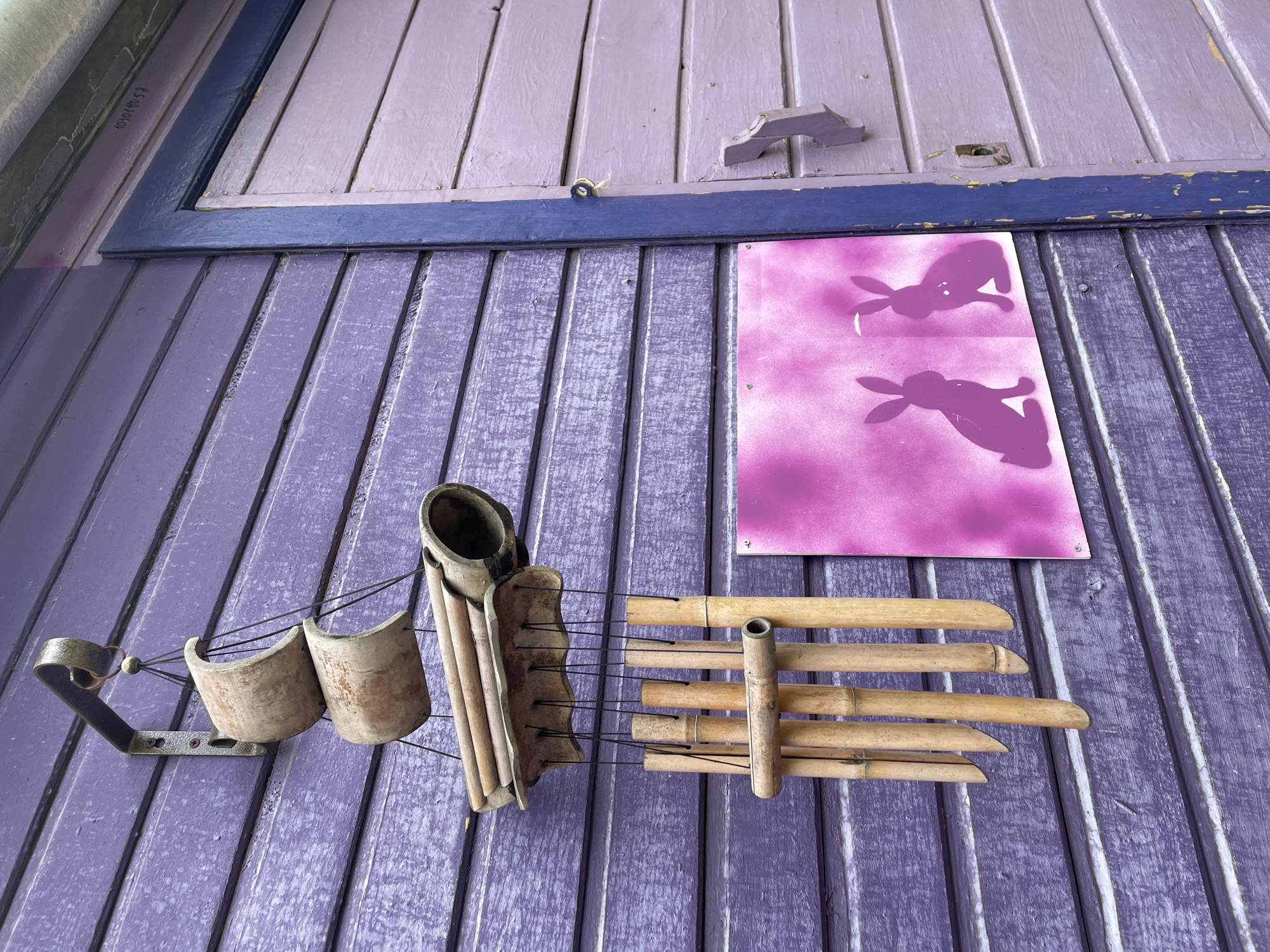 A medium, angled shot shows an exterior wooden wall, painted lavender. The upper portion is a sectioned, smaller planked area with a darker purple border. A lavender sign with two purple rabbit silhouettes is affixed to the wall. Below and to the left of the sign, a metal and clay instrument hangs. To the right of this is a bamboo instrument. The image appears to be taken in natural light. There is no indication in the image that it is located in Khimki, Russia.