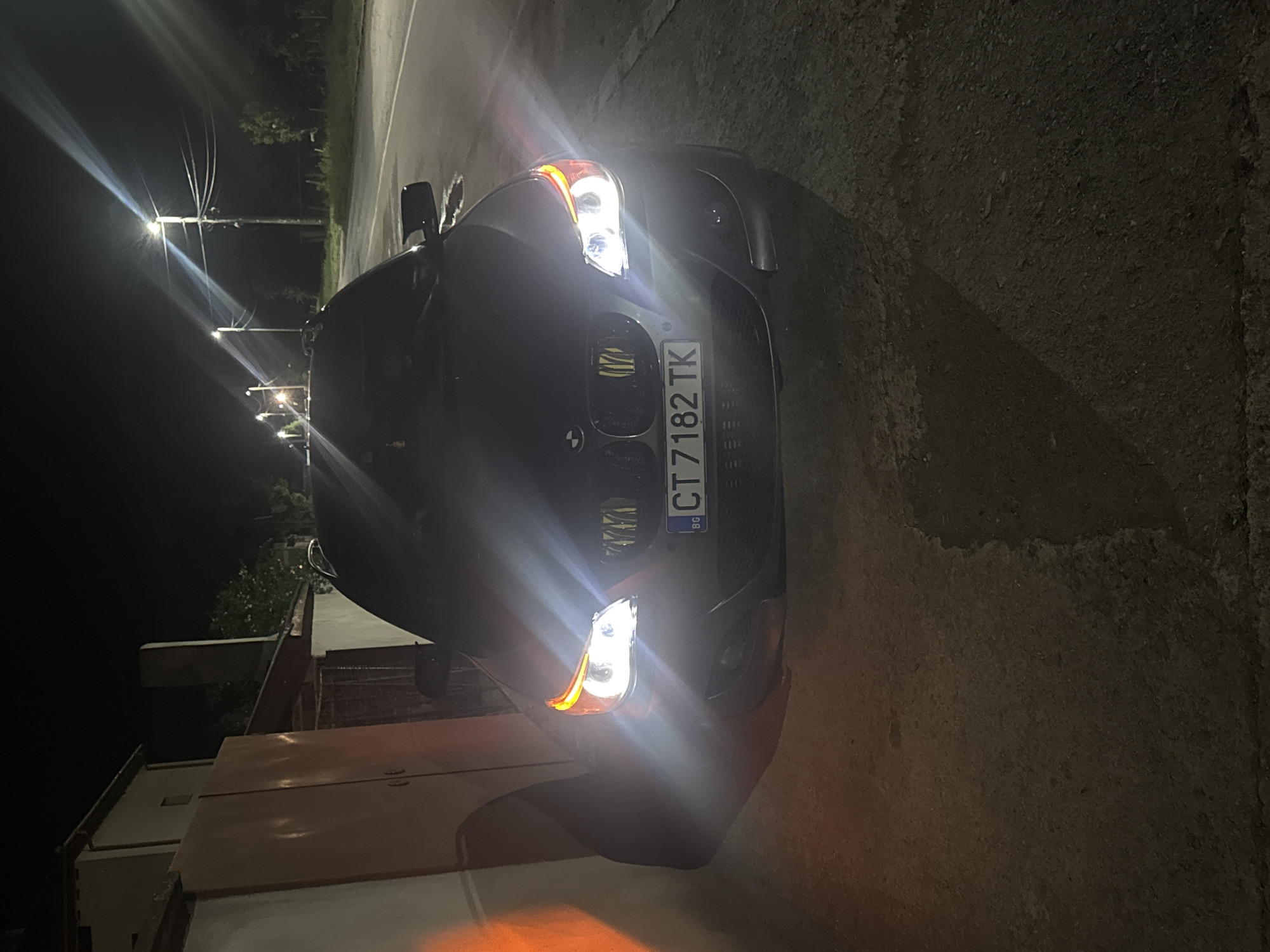 The image is a nighttime shot of a dark-colored BMW with a license plate that reads "CT 7182 TK". The license plate indicates the vehicle is registered in Bulgaria. The car is parked on an asphalt road, and the lighting suggests a city environment. Streetlights are visible in the background, illuminating the scene. The photo's composition and the presence of streetlights suggest the location is likely in a residential area within Sarnevo, Bulgaria. Other objects include an unknown house.