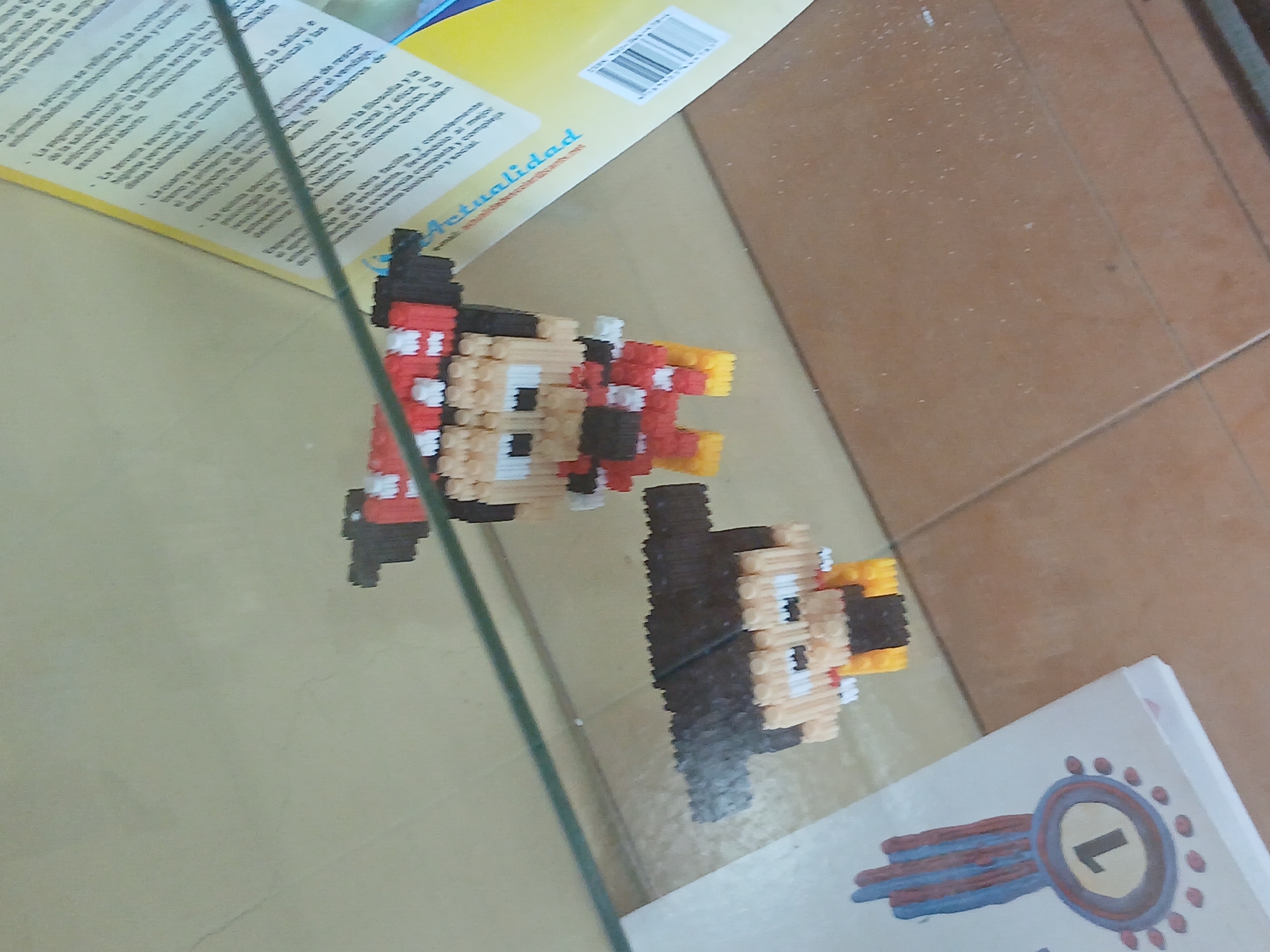 The image shows a shelf with two pixel art figures, possibly characters like Mickey Mouse, displayed behind a glass barrier. One figure is red and white with black accents, and the other is brown, black, and yellow. To the left of the figures is a white booklet with a circular emblem containing the number "1" in black. In the background, a yellow booklet with text in Spanish is visible, possibly a publication or informational material. The environment appears to be an indoor setting, possibly a museum or a display area, with a tiled floor visible beneath the shelf. The overall impression is of a curated exhibit. Without further context or visual cues, the specific location within Juan Griego, Venezuela cannot be determined from the image alone.