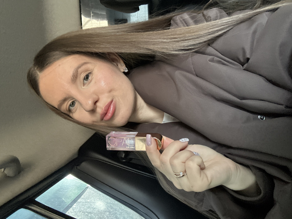 An adult female is depicted in a close-up, chest-up shot, seated inside a vehicle in the city of Zavidovo, Russia. She possesses long, straight light-brown hair and is looking directly at the camera with a subtle smile, her lips appearing glossy. She wears a dark brown, possibly padded, jacket over a lighter garment. Her left hand is raised, holding a transparent, rectangular bottle labeled "OPAL LIP OIL" with a gold-colored base and cap. Her left ring finger displays a two-banded ring with a clear stone. Her fingernails are manicured with light lavender polish featuring white stripe patterns. A small, heart-shaped stud earring with clear stones is visible in her right ear. The interior of the vehicle shows dark upholstery, and an out-of-focus view through a side window suggests an overcast or wet exterior environment.