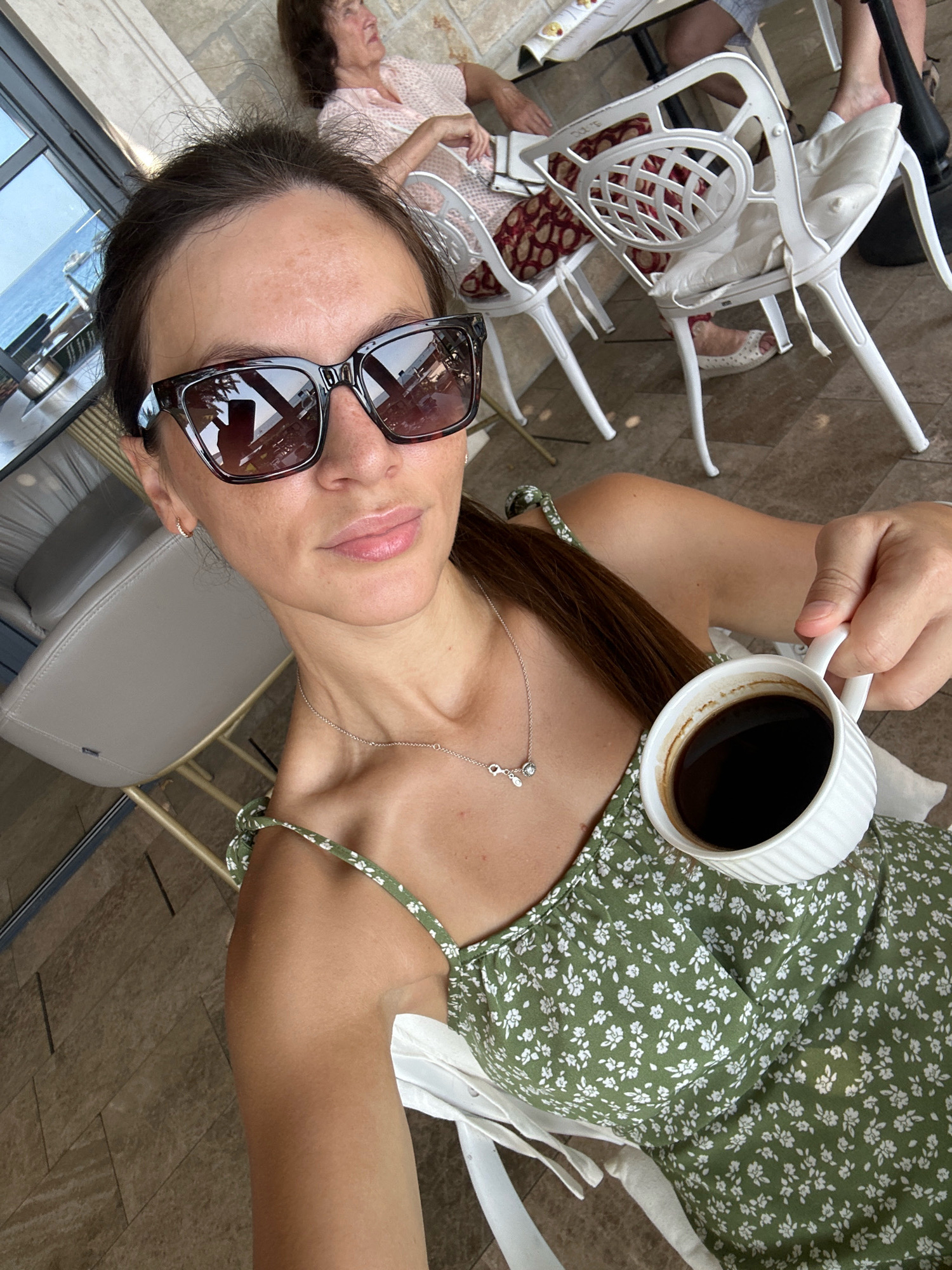 A woman with dark hair, wearing oversized sunglasses and a green floral dress, is holding a white mug of coffee. She is seated at an outdoor cafe with a view of the sea. In the background, other people are seated at tables. The cafe appears to be located in Rafailovići, Montenegro. The overall atmosphere is relaxed and casual, suggesting a leisurely moment.
