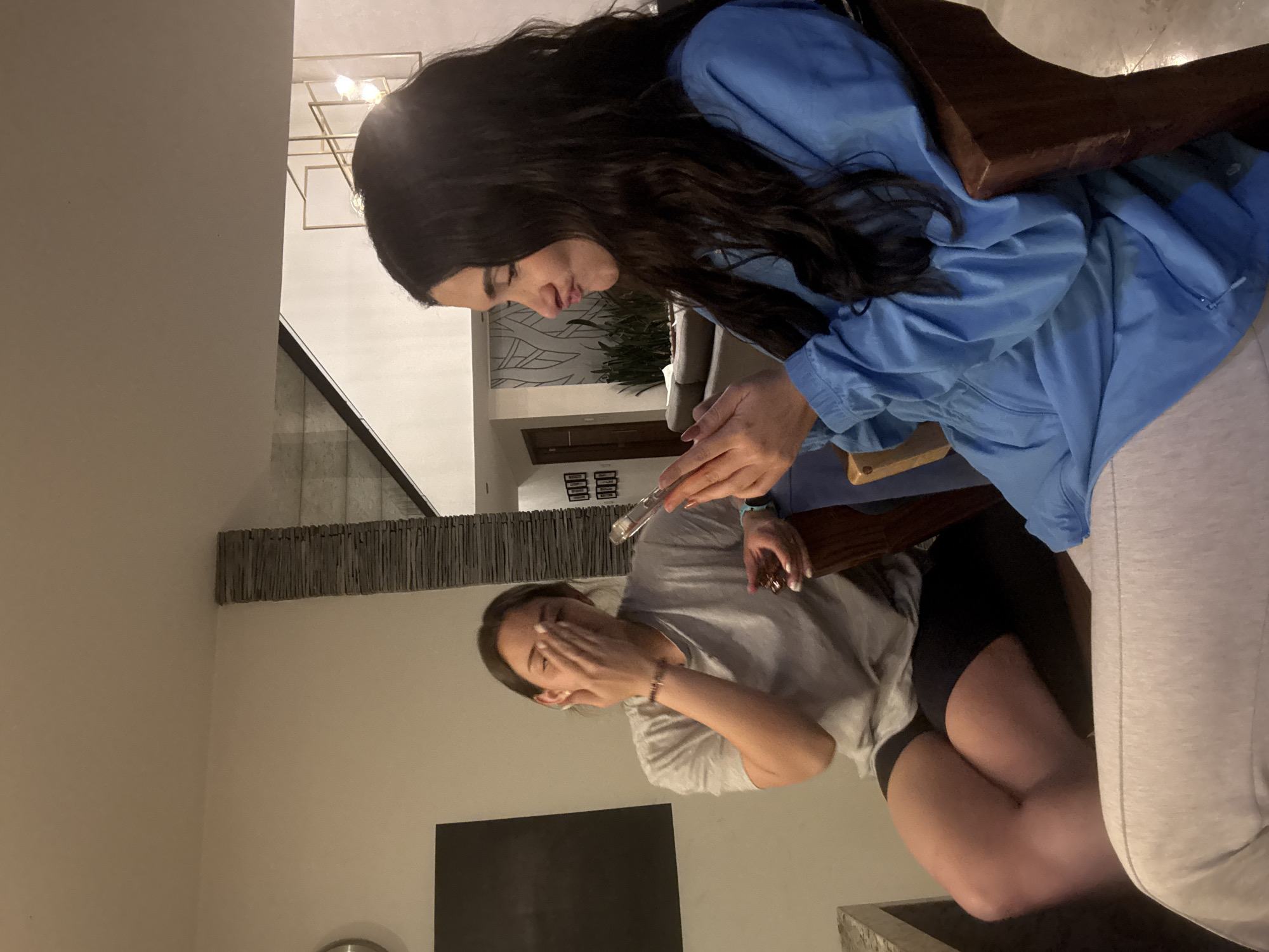 This indoor image, taken in San Bernardino Tlaxcalancingo, Mexico, features two women seated in what appears to be a modern home, though the photograph is rotated 90 degrees clockwise.
The woman positioned higher in the rotated frame has long, dark hair and is wearing a blue jacket over light-colored bottoms. She is looking downwards with a focused expression, holding a clear, slender object, possibly a glass tube or vape pen, with both hands. She appears to be interacting with or preparing something with this object.
Seated lower in the frame is another woman with shorter dark hair, dressed in a grey t-shirt and black shorts. Her left hand is raised and covers part of her face, suggesting amusement, shyness, or a strong reaction. A beaded bracelet is visible on her left wrist. Both women are sitting on chairs with visible wooden elements.
The background reveals contemporary interior design elements. A distinctive light fixture with a geometric, golden-toned frame hangs from the ceiling. A textured wall feature made of horizontal slats, possibly wood or stone, runs across the mid-background. Behind the second woman, a black rectangular object, likely a television screen or framed art, is mounted on the wall. Green plants are visible further back, adding to the indoor ambiance. The lighting suggests artificial sources, indicating it might be evening or night. There is no visible text in the image.
