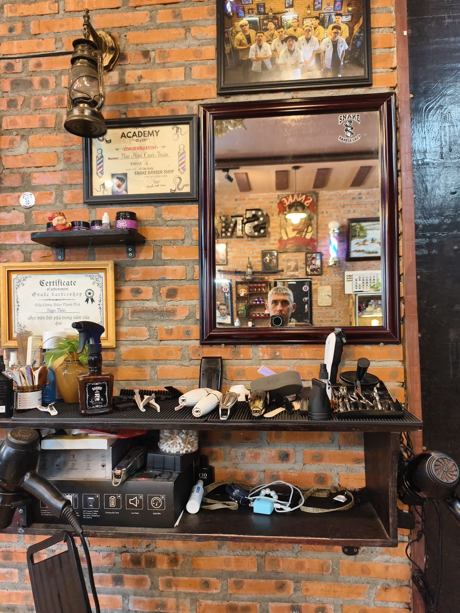 The image captures a wall of exposed brick in what appears to be a barbershop in Bac Nha Trang Ward, Vietnam. On the left, a vintage oil lamp hangs from a bracket. Below it, a framed certificate from "ACADEMY SNAKE BARBER SHOP" is displayed. A narrow shelf above holds a few small jars and figurines.
A large, dark-framed mirror dominates the center of the image, reflecting the interior of the shop. A man's face is visible in the reflection, looking directly at the camera. The mirror is adorned with a "SNAKE BARBER SHOP" sticker. Various items are arranged on shelves below the mirror.
The top shelf holds numerous hair styling tools and accessories, including scissors, clippers, combs, and brushes, neatly organized. A bottle of "BARBER" branded product sits in the center. A plant in a vase is also visible.
The shelf below displays more barbering equipment such as hair clippers and trimmers, along with boxes and bottles. A black hairdryer is prominently placed on the left side of this shelf.
The lower shelf, made of dark wood, shows more personal items and accessories, including a small blue electronic device, some jewelry or beads, and a coiled cable. A second black hairdryer is visible on the right side of the image, attached to the wall. The overall environment suggests a well-equipped and possibly traditional barbershop.