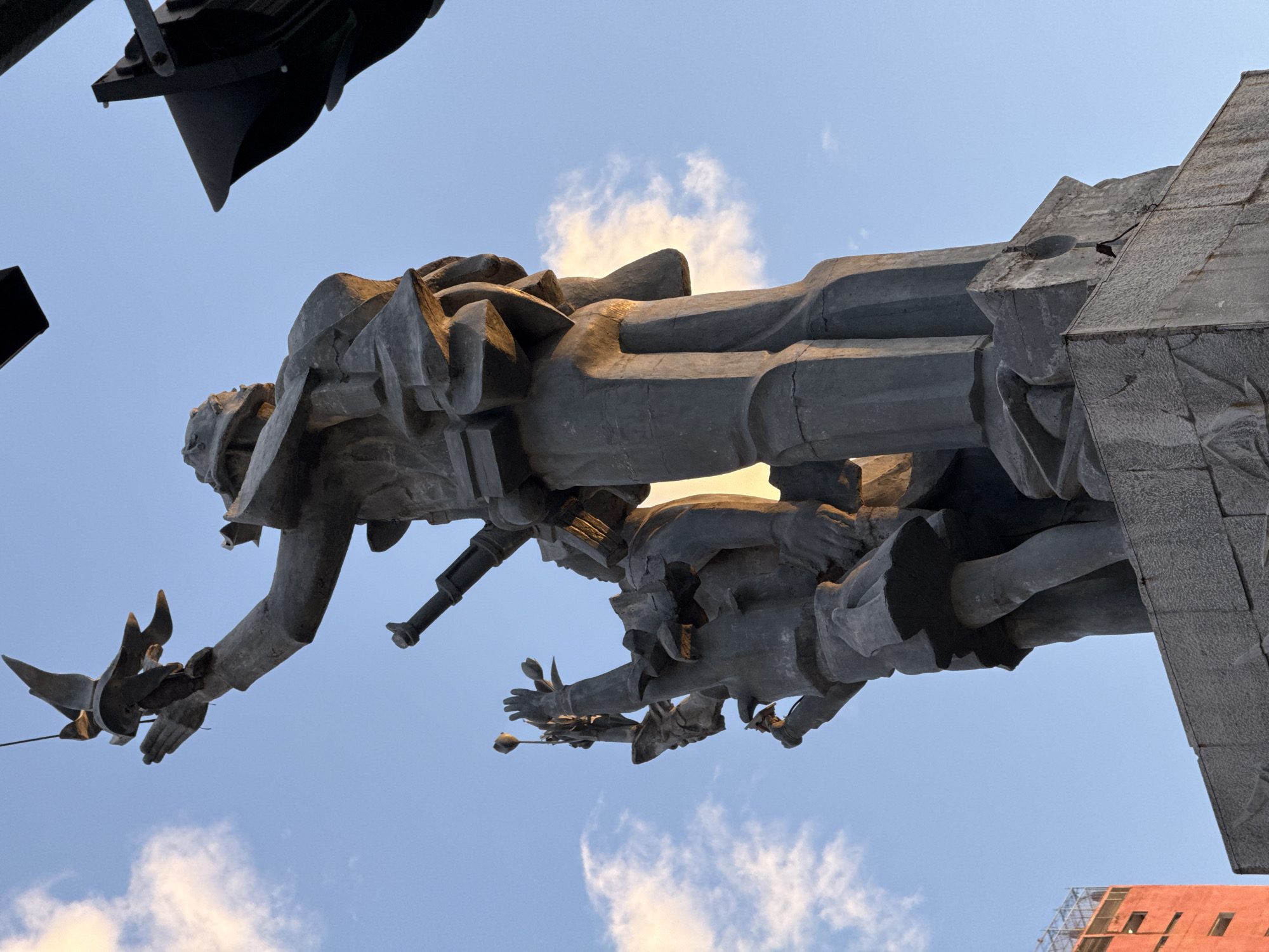 This image shows a low-angle view of a monumental stone sculpture against a bright blue sky with scattered clouds. The sculpture depicts several figures, appearing to be heroic in stature. The primary figure in the foreground is a large, imposing male figure holding a bird in its outstretched hand. To the left and slightly behind this figure is a smaller figure, possibly a child or a woman, reaching upwards with flowers. The figures are rendered in a stylized, modernist aesthetic with sharp angles and simplified forms.
In the background, to the left, a portion of a tall, orange building with visible windows and scaffolding is visible. Overhead, black objects, possibly lights or parts of a structure, are partially visible in the upper portion of the frame. The overall impression is that of a memorial or monument, set against an urban backdrop.
This monument is the **Monument to Vietnamese Heroic Mothers** located in **Nha Trang, Vietnam**. It commemorates the women of Vietnam who suffered greatly during the wars. The main figure represents a mother, and the bird symbolizes peace or a message being sent. The smaller figure is likely a child.
