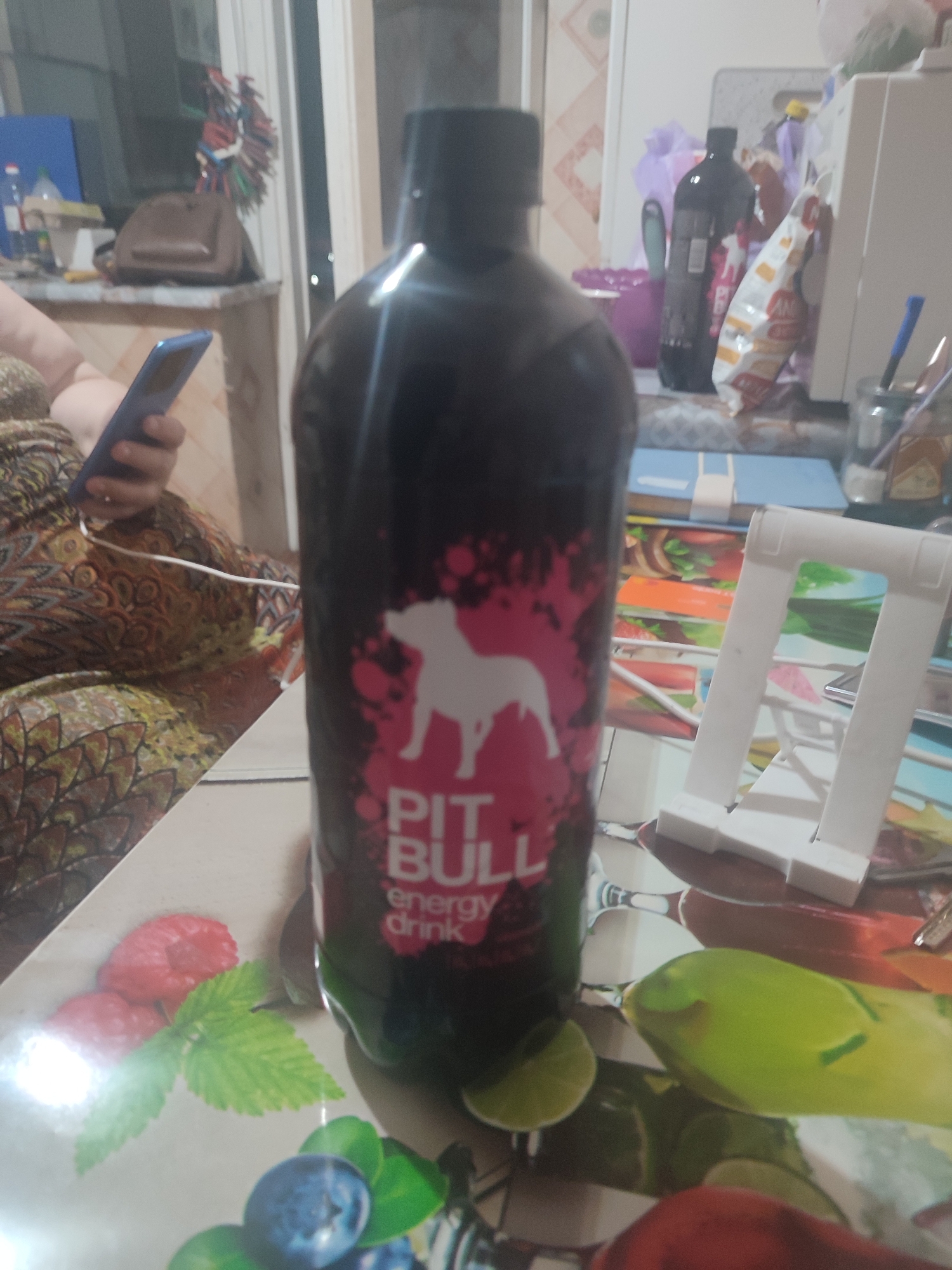 This image depicts an indoor scene, likely a home or office environment. In the foreground, a large, dark-colored bottle of "PIT BULL ENERGY DRINK" is prominently displayed. The bottle has a pink graphic featuring a white silhouette of a pit bull dog. To the left of the bottle, a person is partially visible, seated and holding a blue smartphone. Their clothing is a patterned fabric with earthy tones.
The background reveals a cluttered surface with various items, including a white tablet stand, a blue book, and what appears to be packaging. Further in the background, a purse or bag is visible on a shelf, along with other indistinct objects. The lighting suggests an interior setting, possibly near a window or artificial light source. The overall impression is a casual, everyday moment captured in a domestic space. The specific location in Chernivtsi, Ukraine, cannot be determined from the visual information provided in the image.