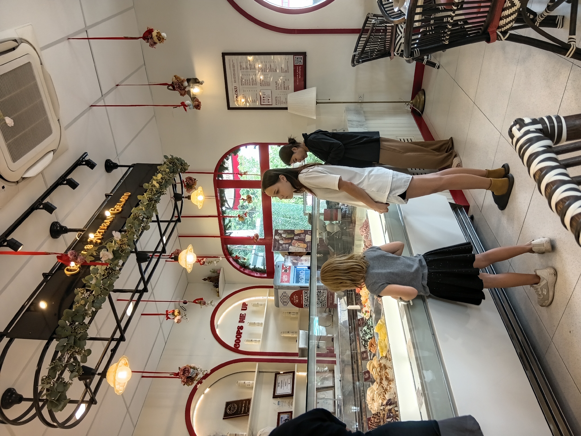The image depicts the interior of an ice cream shop. The shop is decorated with hanging garlands of greenery and flowers, as well as festive red ribbons and small ornaments suspended from the ceiling. There are several display cases filled with various flavors of gelato. Three people are visible: a young girl in a grey t-shirt and black skirt is standing in front of a display case, looking at the gelato. To her right, two young women are standing. One, wearing a white t-shirt and shorts, is looking towards the display case. The other, in a black jacket and brown pants, is looking down at her phone. In the background, a menu board is displayed on the wall, and there are chairs and tables, suggesting a seating area. The lighting appears to be a mix of overhead lights and decorative lamps. The shop is clean and well-lit. There is no specific indication within the image to confirm the location as Phra Nakhon Si Ayutthaya City Municipality, Thailand.