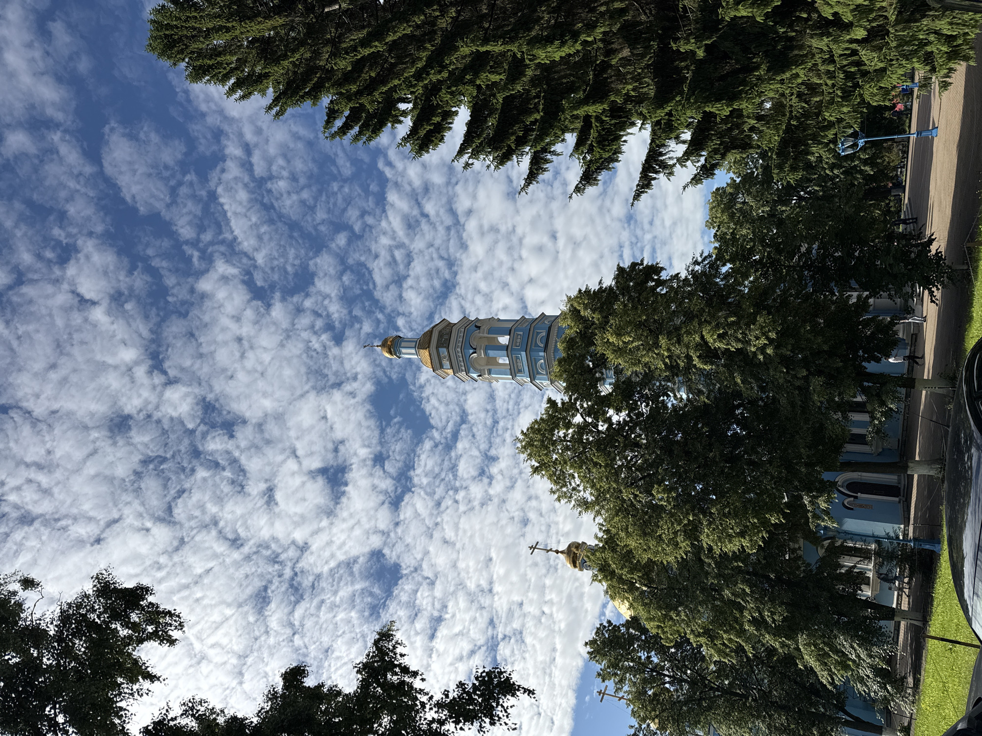 A low-angle outdoor shot shows a blue and gold Orthodox church with a tall, slender bell tower piercing a sky filled with scattered white clouds against a bright blue backdrop. The church building, painted a vibrant blue with white trim, has golden domes visible amongst the dense green foliage of several large trees that frame the scene. The bell tower, also blue and gold, features arched openings and a cross atop its spire. To the right, a large, dark green evergreen tree with thick, drooping branches partially obscures the sky. The ground in front of the church is paved, with a green lawn visible to the left and a pathway leading into the distance where several individuals are walking.