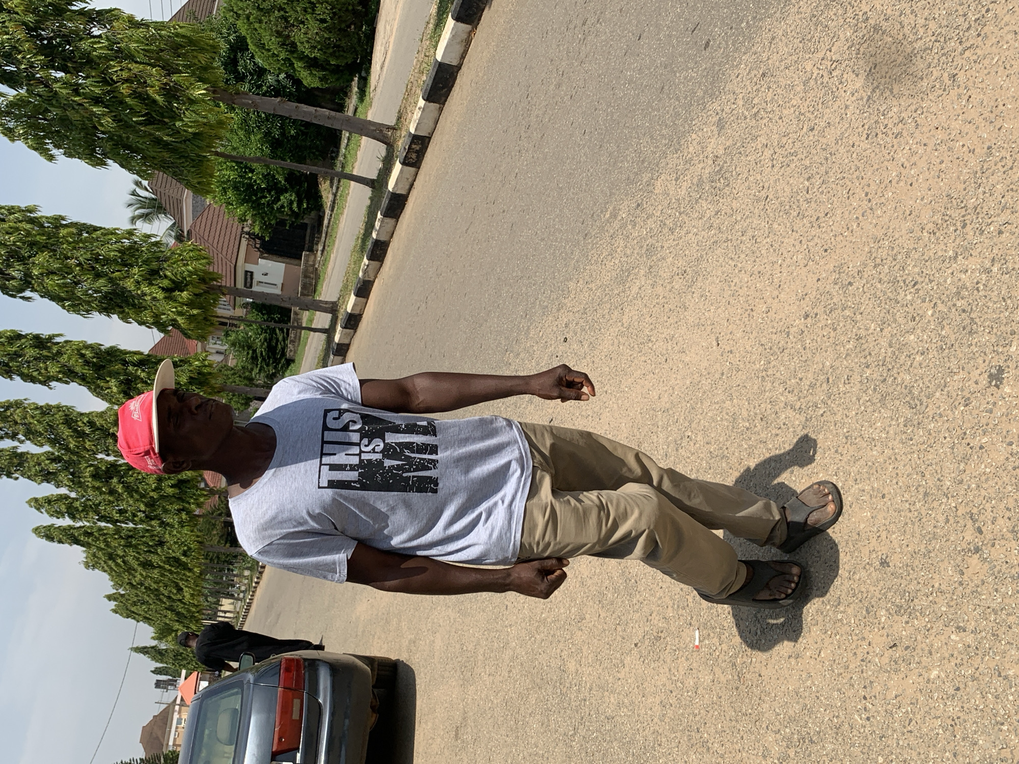 A man wearing a red baseball cap, a gray t-shirt with the text "THIS IS MY" printed in black, and beige pants walks down a paved road in Abuja, Nigeria. He is wearing dark flip-flops. A dark-colored sedan is parked on the left side of the road, and behind it, another person in dark clothing is walking away from the camera. The road is lined with tall, thin trees and houses with brown roofs in the background. The sky is clear and bright, suggesting a sunny day. The scene appears to be in a residential or suburban area.