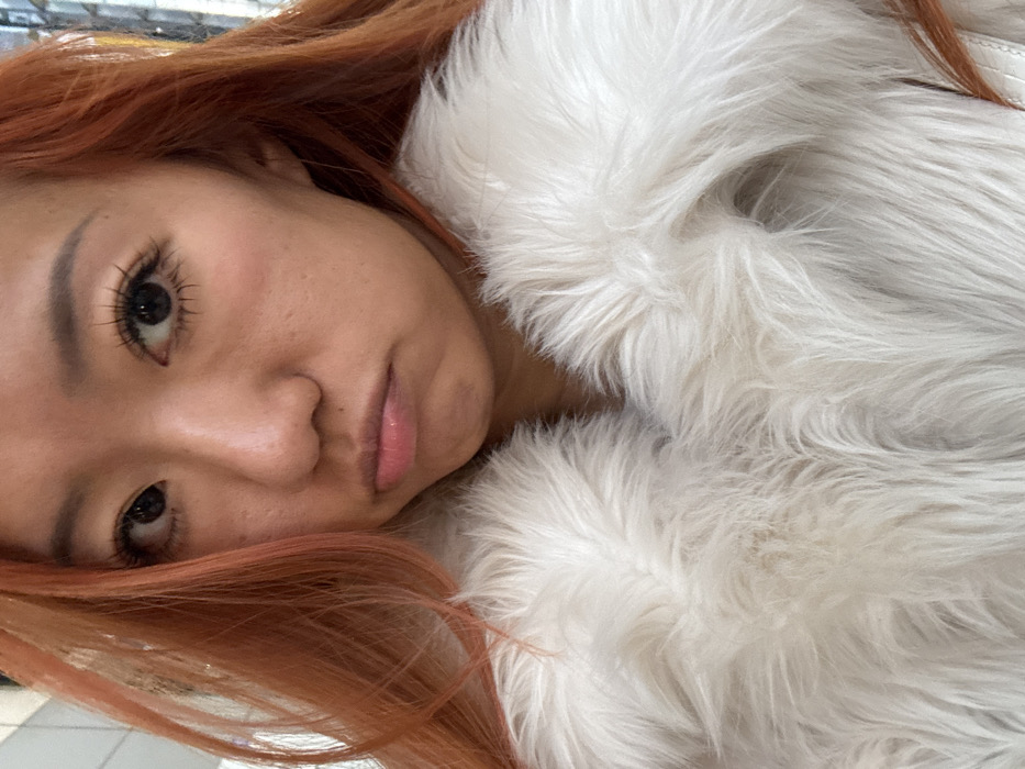 A woman with orange-red hair poses close to the camera. She has dark eyes, fair skin, and full lips. She appears to be wearing heavy mascara. Her head is tilted, and she rests against a fluffy, white faux fur coat or wrap. The background is indistinct and slightly blurred, but a glimpse of a white tiled floor suggests an indoor setting. The lighting is soft, highlighting the textures of the hair and fur.