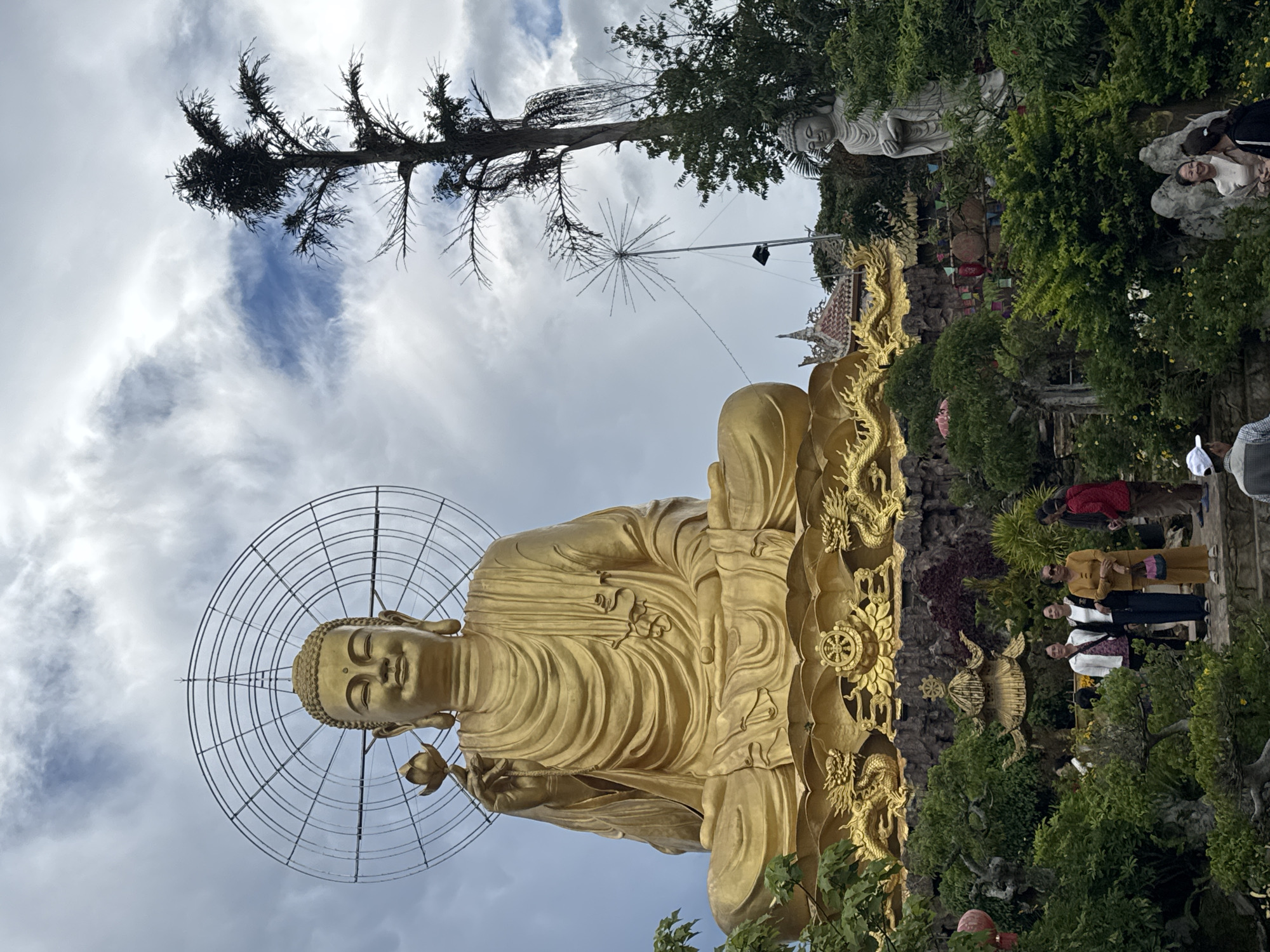 In Lâm Viên - Đà Lạt, Vietnam, a colossal golden Buddha statue sits in meditation, holding a lotus flower. The statue, approximately 50 feet tall, is adorned with a halo and rests on a gilded base featuring dragon motifs. Behind the Buddha, a tall, slender tree with sparse foliage stretches towards a cloudy sky. To the right, a smaller, white Buddha statue stands amidst lush greenery. The foreground is a vibrant garden with meticulously pruned trees and decorative structures. Several individuals, appearing to be tourists, are scattered throughout the garden, some posing for photos, others observing the statues, and a few engaging in conversation. The atmosphere is serene, suggesting a place of worship or a significant tourist attraction.