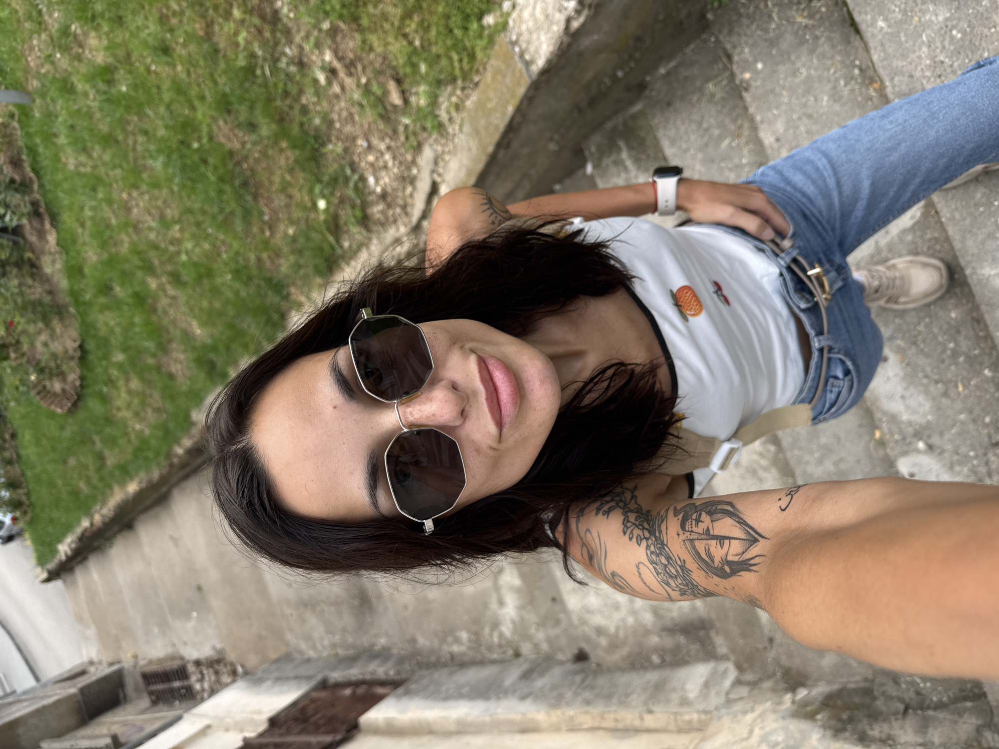 A young adult female is depicted in a high-angle selfie, positioned outdoors on a descending concrete staircase. She has dark hair, wears hexagonal framed sunglasses that obscure her eyes, and exhibits freckles across her nose. Her attire consists of a white tank top with an orange fruit emblem on the front, light blue denim jeans, and light-colored sneakers. A thin brown belt with a gold buckle is visible at her waist, and a white smartwatch with a red stripe on the band is worn on her right wrist. Multiple tattoos are present: a prominent design featuring a split face (half human, half stylized horned figure) encircled by foliage on her left upper arm, and a sunburst pattern on her right upper arm. To the left of the subject, an aged, light-colored building or wall with visible brown shutters stands. To her right, a grassy area with some dry patches is bordered by a low concrete barrier. A road and faint background elements are visible in the distance. The scene is situated in the city of Pleven, Bulgaria.