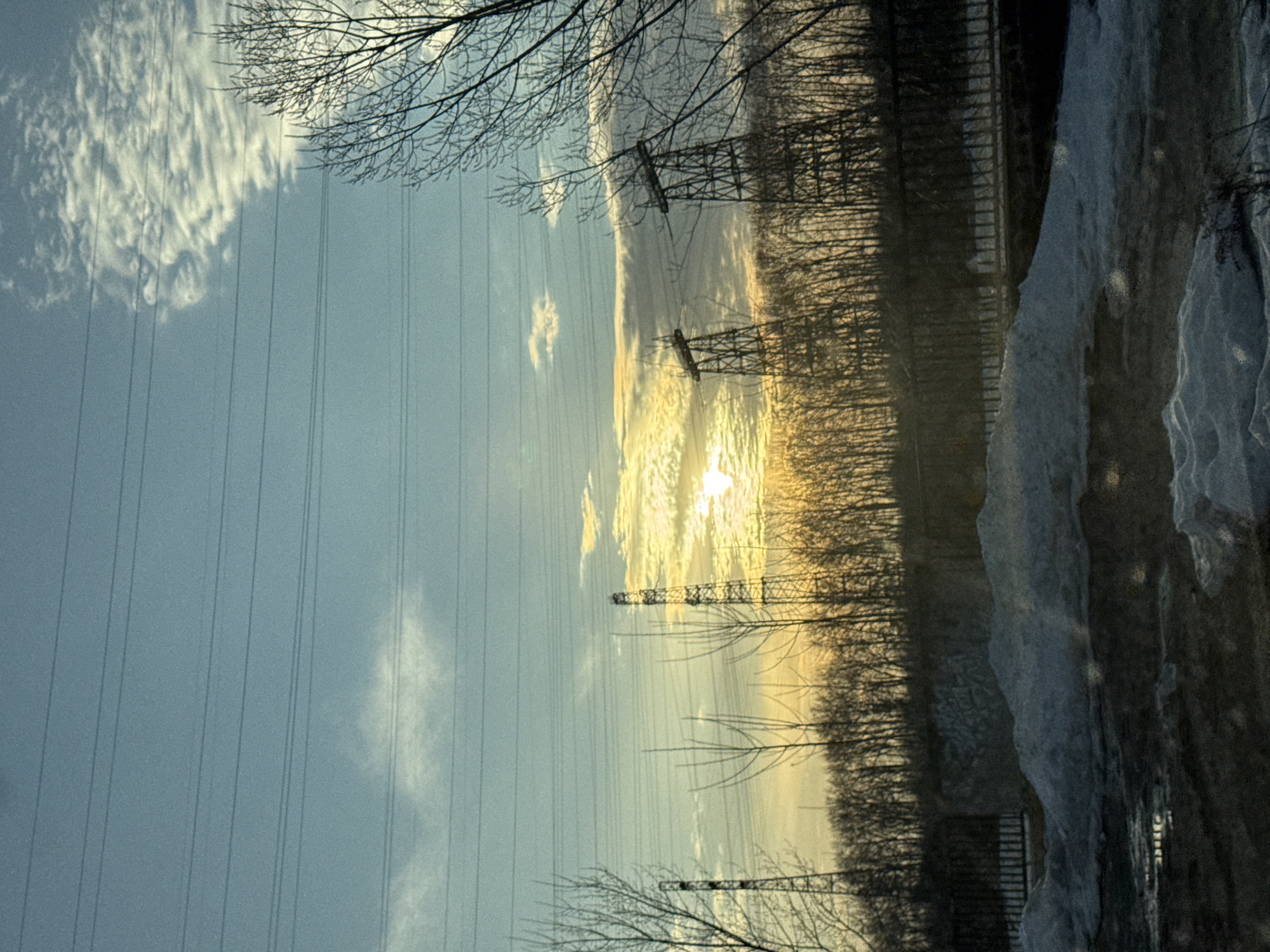 The image shows a winter scene in what could be Chaikovsky, Russia, based on the presence of elements often found in such areas. The sky is predominantly blue, with some patches of white clouds in the upper left. Power lines stretch across the sky. The sun is visible, blurred through clouds. Silhouettes of bare trees are present in the upper part. Some snow or ice features are on the right edge of the image. Transmission towers are partially visible amid trees. The image appears to be rotated clockwise.