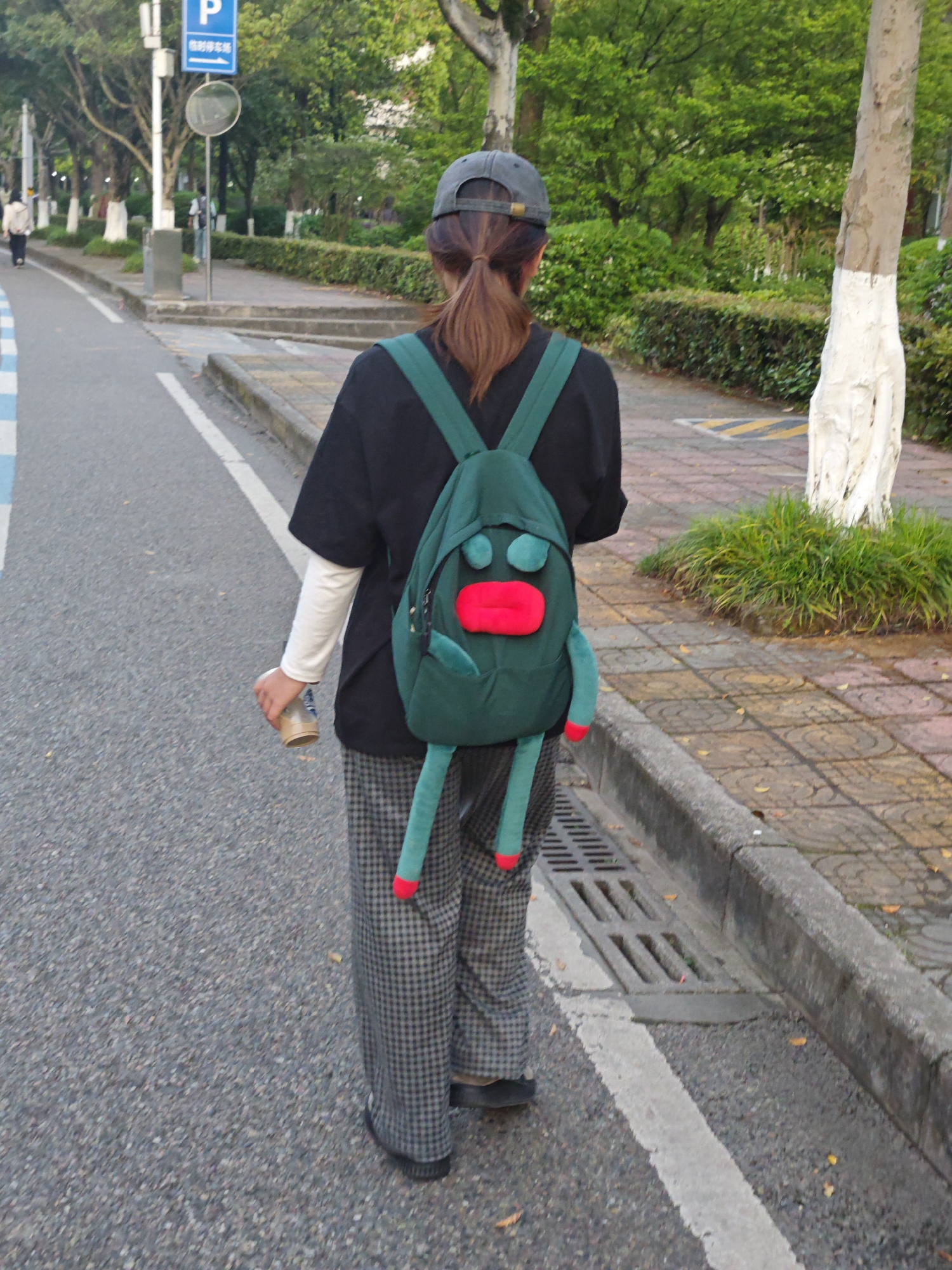 A person is walking away from the camera on a road in what appears to be an urban area in Chongqing High-tech Zone, China. The individual is wearing a dark-colored t-shirt over a long-sleeved white shirt, plaid pants, and a cap. They are carrying a beverage container in their left hand and have a green backpack with a cartoonish face design. The road is lined with trees and foliage, and there is a sidewalk to the right with a curb and a drain. Street signs are visible in the background.