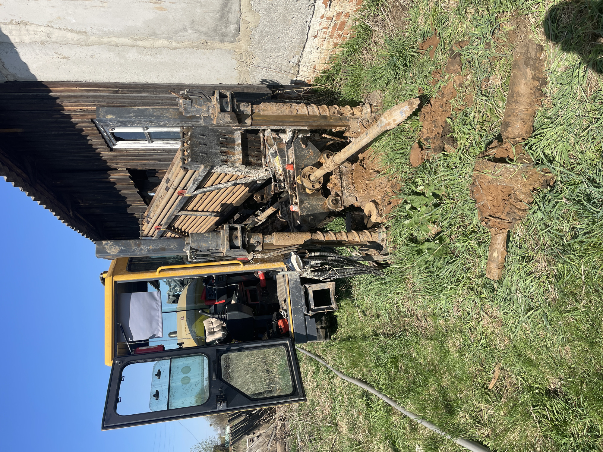 A compact drilling rig is positioned in a grassy area next to a building with a wooden facade and a brick foundation, possibly in a rural or semi-rural setting. The building features a window with a white frame. The drilling rig is yellow and black, with an open cabin and an extended drilling arm. A section of the drilling pipe lies on the ground, covered in mud. The immediate environment consists of grass and disturbed earth. The sky is clear and blue. The location is Поедуги, Russia.