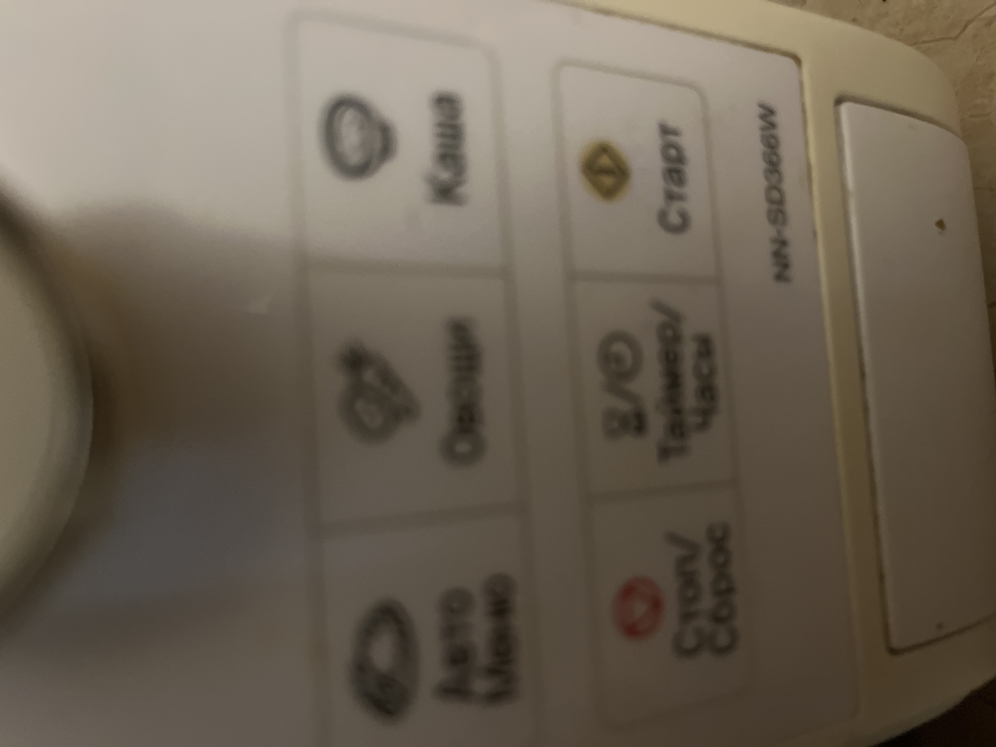 The image shows a close-up of a control panel on a white appliance, likely a rice cooker. The panel has several buttons with icons and text in Russian. From top to bottom and left to right, the visible buttons include:
* A circular icon with a swirl pattern and the Russian word "Каша" (Kasha), likely indicating a porridge or cereal setting.
* A button with a yellow play/start symbol and the Russian word "Старт" (Start).
* A button with an icon resembling a pot or bowl and the Russian word "Опции" (Options).
* A button with two icons, possibly a clock or timer symbol, and the Russian words "Таймер/ Часы" (Timer/ Hours).
* A button with an icon resembling a plate or bowl and the Russian words "Авто Меню" (Auto Menu).
* A button with a red circle and a triangle inside, and the Russian words "Стоп/ Сброс" (Stop/ Reset).
On the right side of the panel, there is text that reads "NN-SD366W", which is likely the model number of the appliance. The appliance is white and appears to be in good condition, though the image quality is somewhat blurry. The overall setting is indoors, with a neutral background. There are no people visible in the image, and no specific time of day or weather conditions can be inferred.