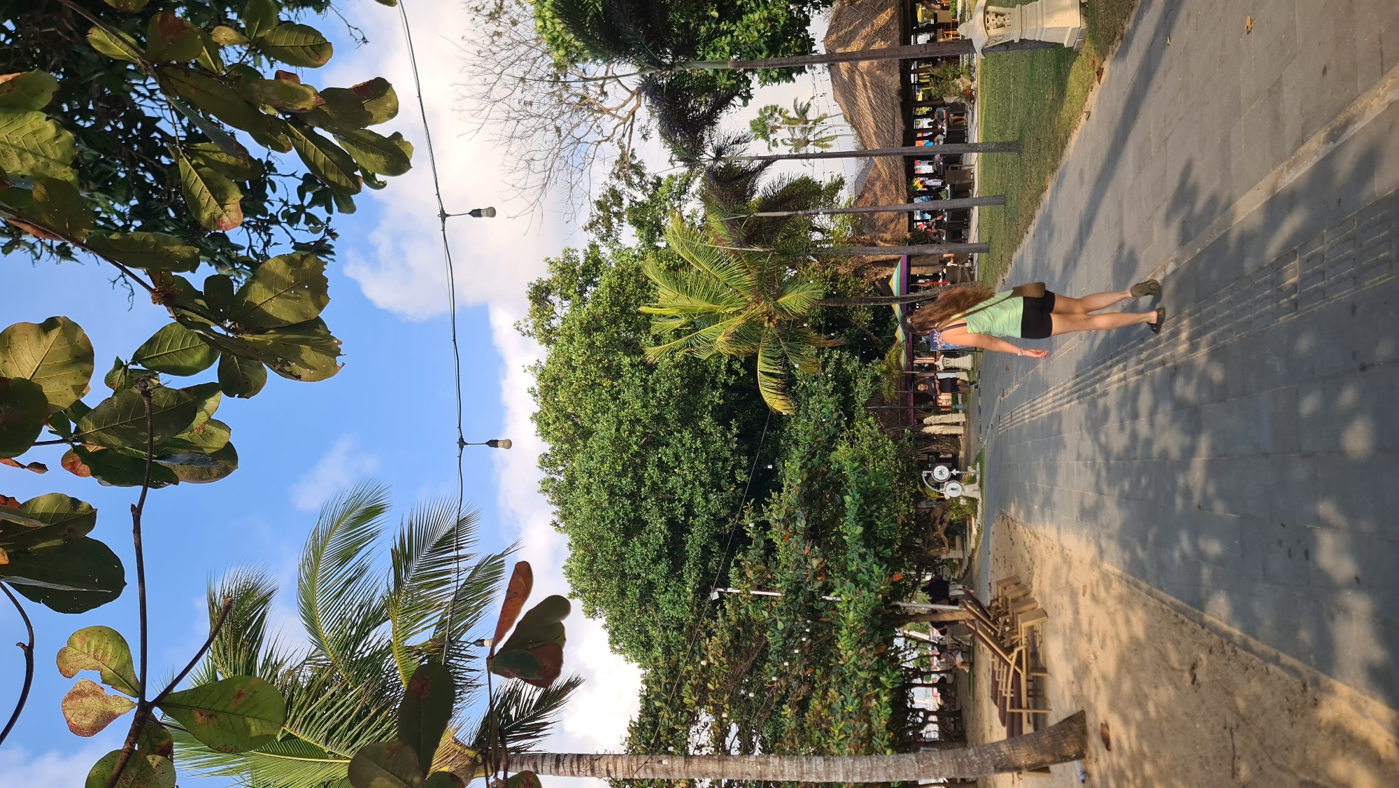 A young woman with long brown hair walks down a paved path in a tropical outdoor setting, possibly a beach resort. She is wearing a light green tank top and black shorts. Behind her, the path is lined with lush green trees and palm trees, with thatched-roof structures and outdoor seating visible in the distance. The ground on the left is sandy, with several empty lounge chairs. String lights are visible hanging overhead. The sky is blue with scattered clouds. The scene appears to be in a warm, sunny climate. This location could be near a beach in Denpasar, Indonesia, given the tropical vegetation and resort-like atmosphere.