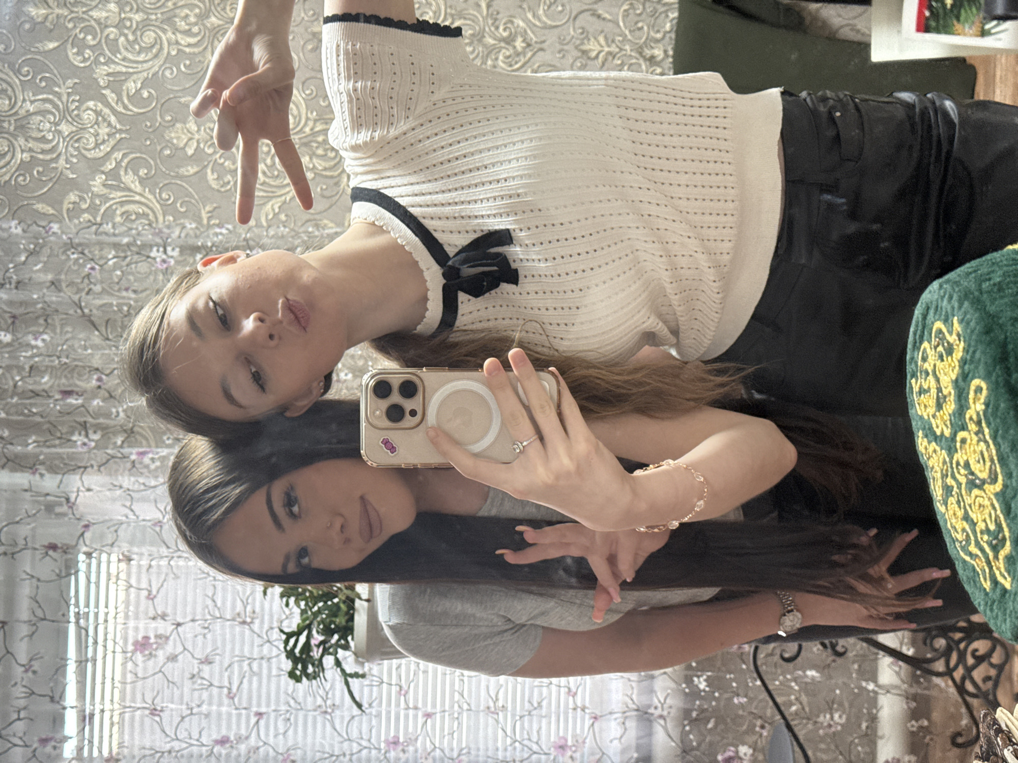 Two young women are taking a selfie in a mirror. One woman, positioned above, has blonde hair and is wearing a white top with black trim and a black bow, and black pants. She is making a peace sign with her hand. The other woman, positioned below, has long dark hair and is wearing a gray shirt. She is holding the phone. The reflection in the mirror shows a room with floral wallpaper and a window with patterned curtains. There is also a plant, a green cushion with gold embroidery, and other furniture visible in the background. The image suggests a casual and informal setting. There is no information suggesting this image was taken in the city of Konovalovka, Russia.
